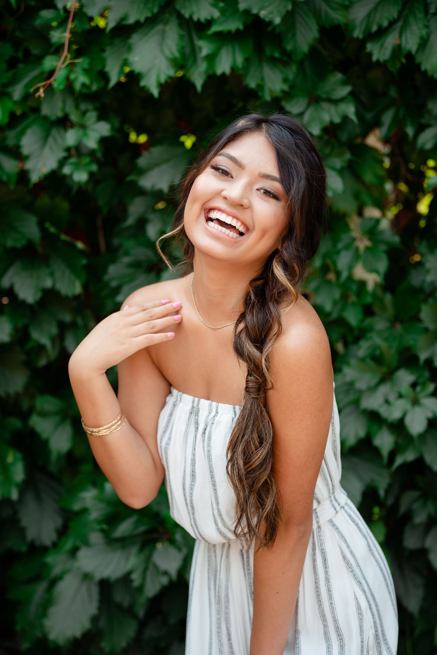 A joyful moment captured outdoors against green foliage wearing a white summer dress with long wavy hair.