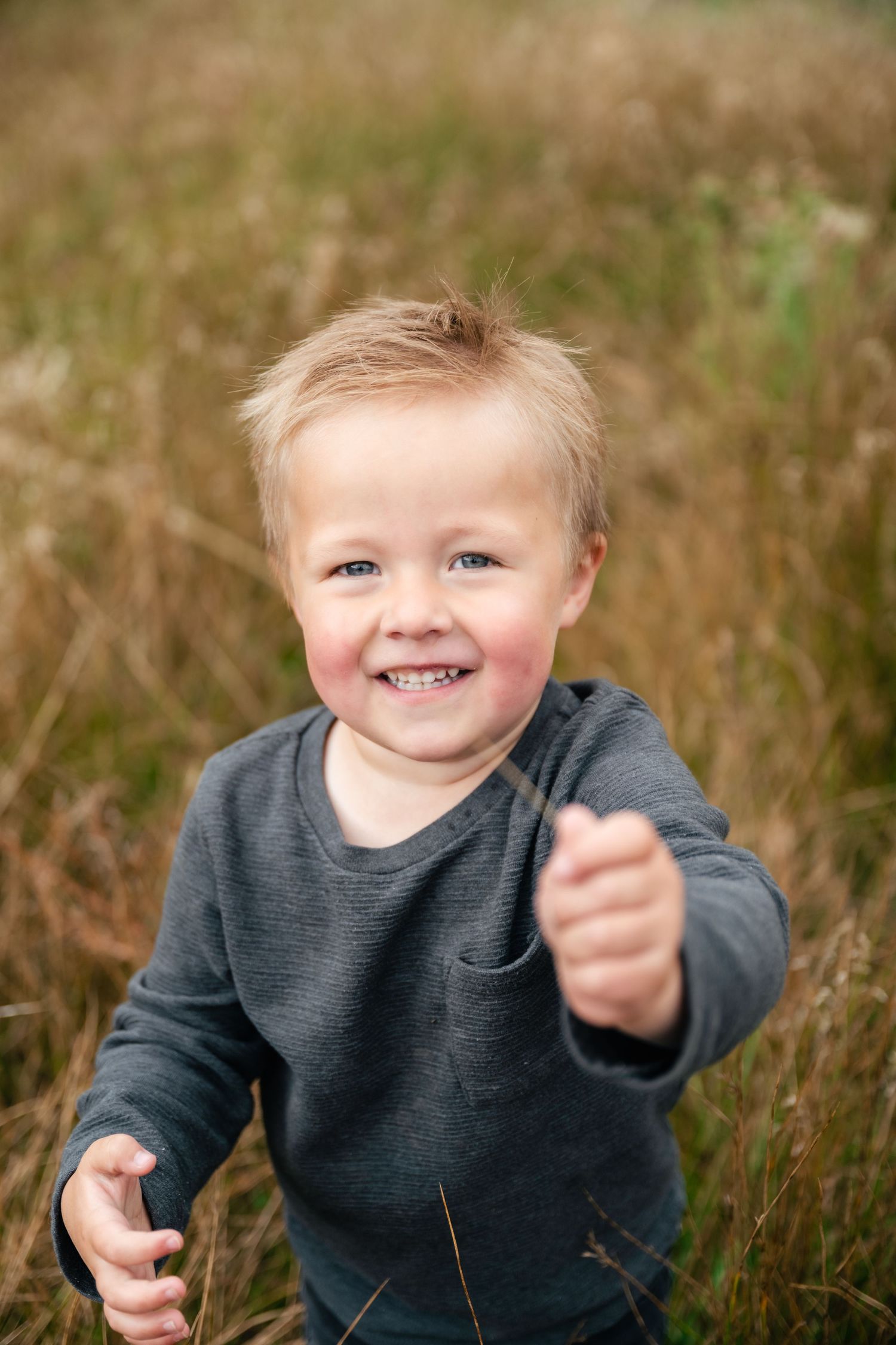 A young child in a gray sweater plays joyfully in a field of tall grass.