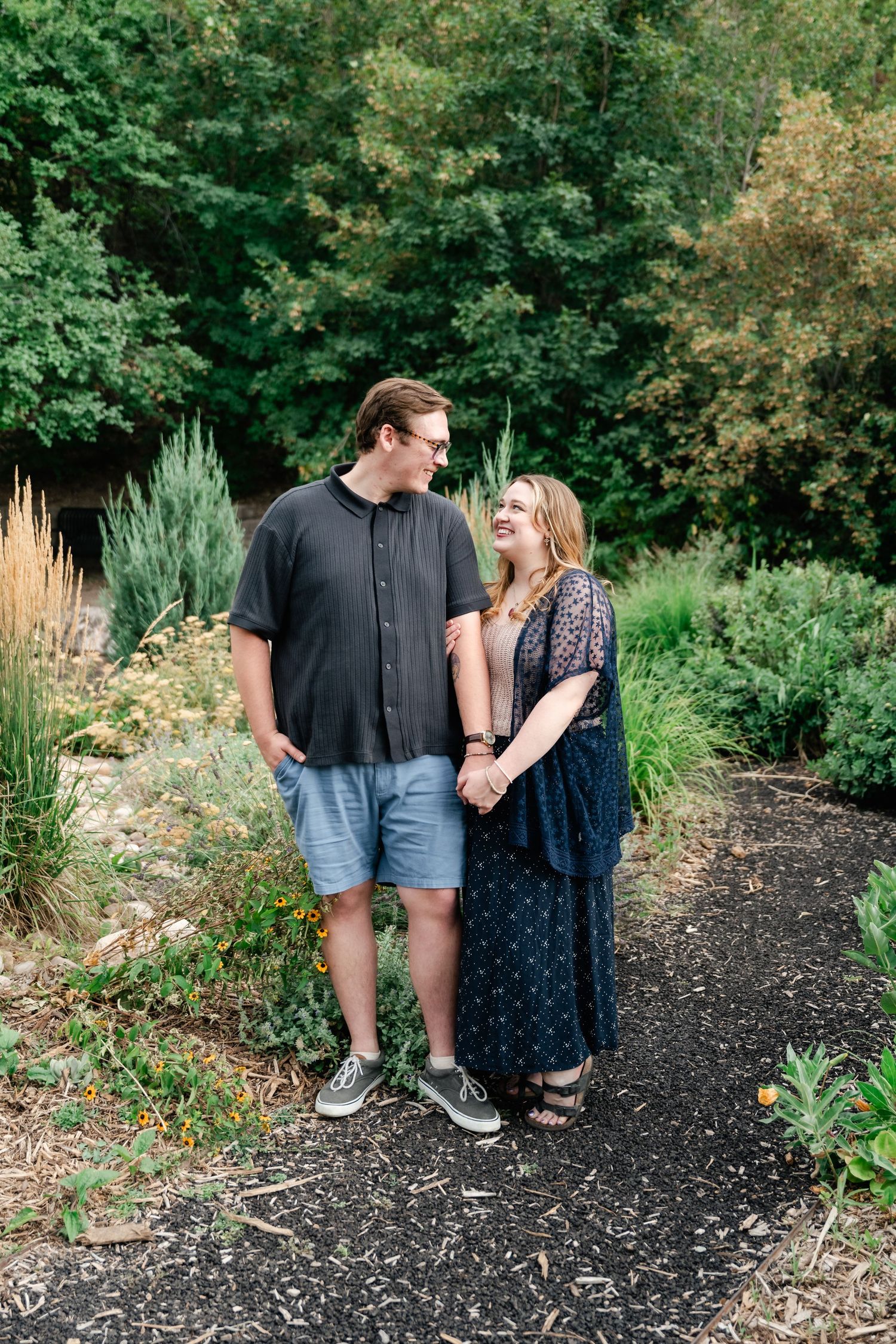 A couple shares a joyful moment together while walking along a garden path surrounded by lush greenery and flowers.