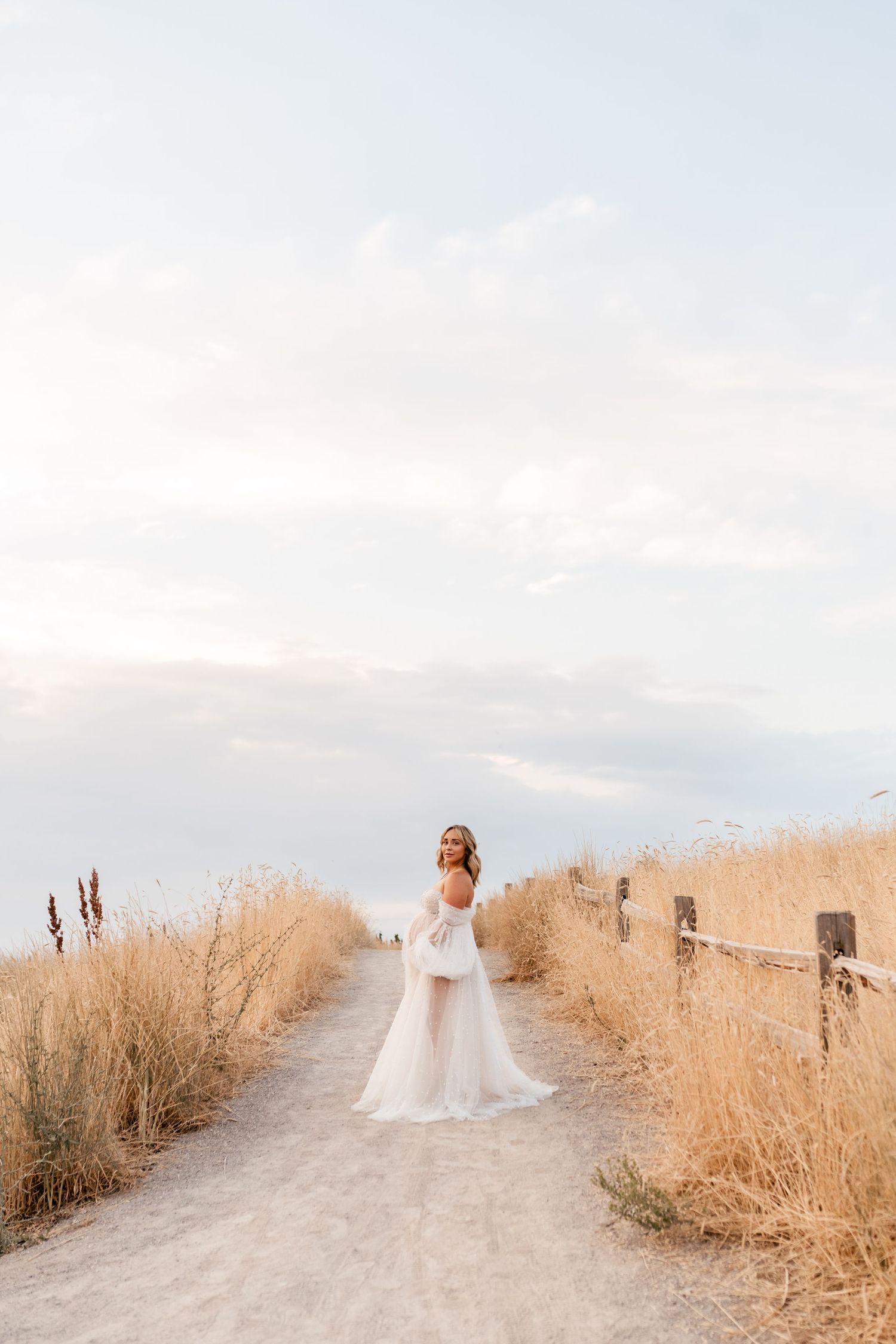 A person in a flowing white dress walks along a winding dirt path through golden dried grass at sunset.