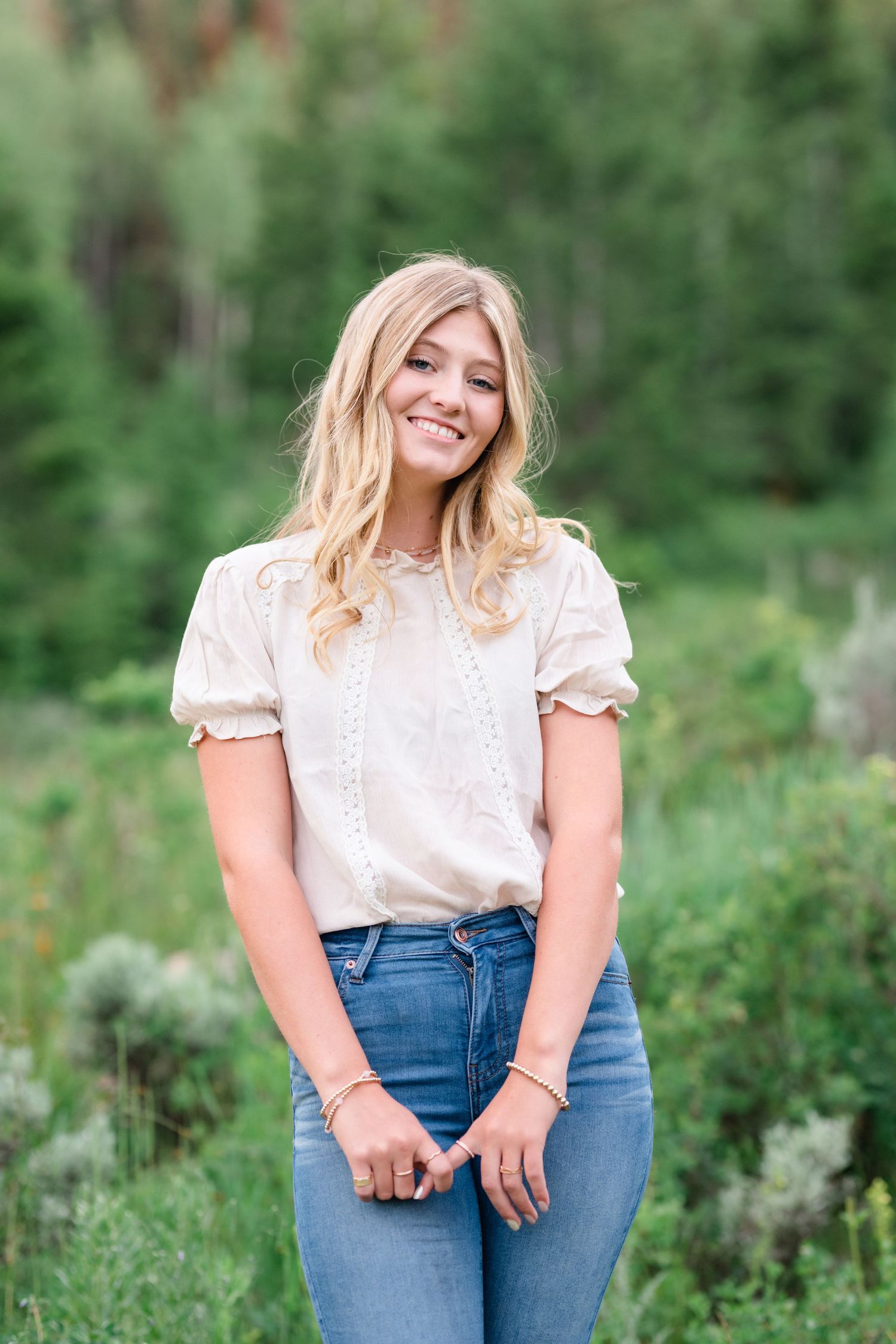 A person in a white blouse and high-waisted jeans poses outdoors against a lush green natural backdrop.