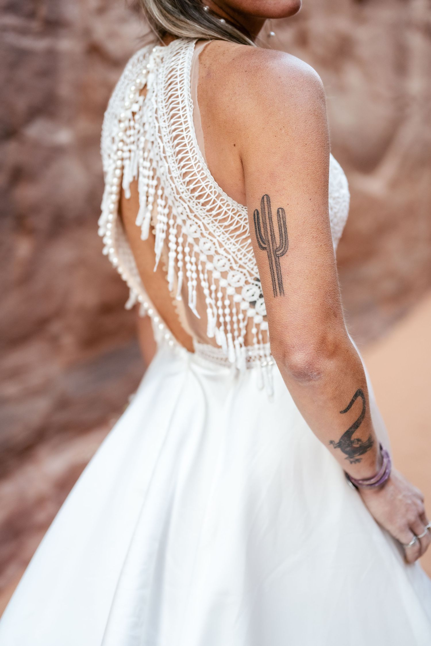 Close-up of a white wedding dress with macrame fringe detail and a cactus tattoo on the wearer's arm.
