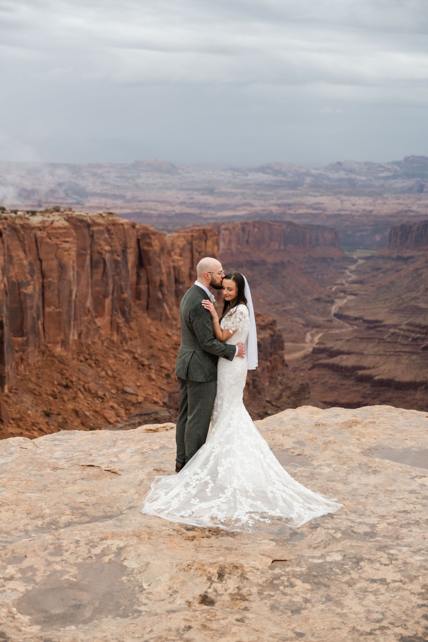 A wedding couple embraces on a scenic desert cliff with vast canyons in the background.
