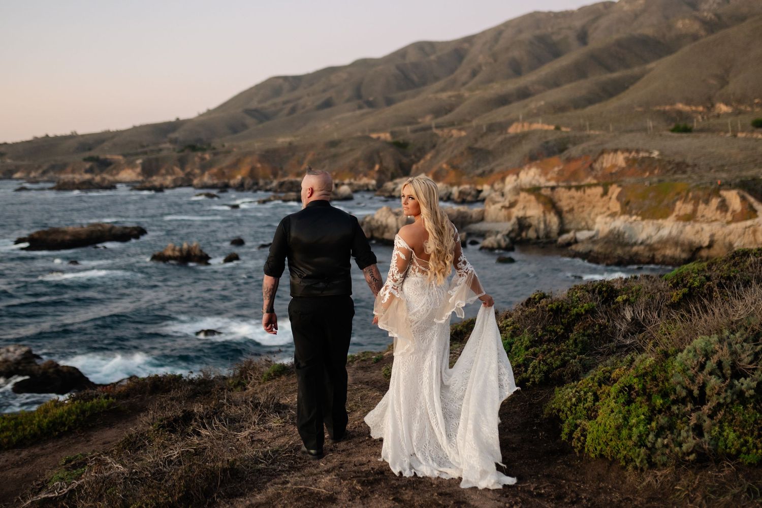 A couple in wedding attire walks along a dramatic Big Sur coastline with rocky cliffs and crashing waves.