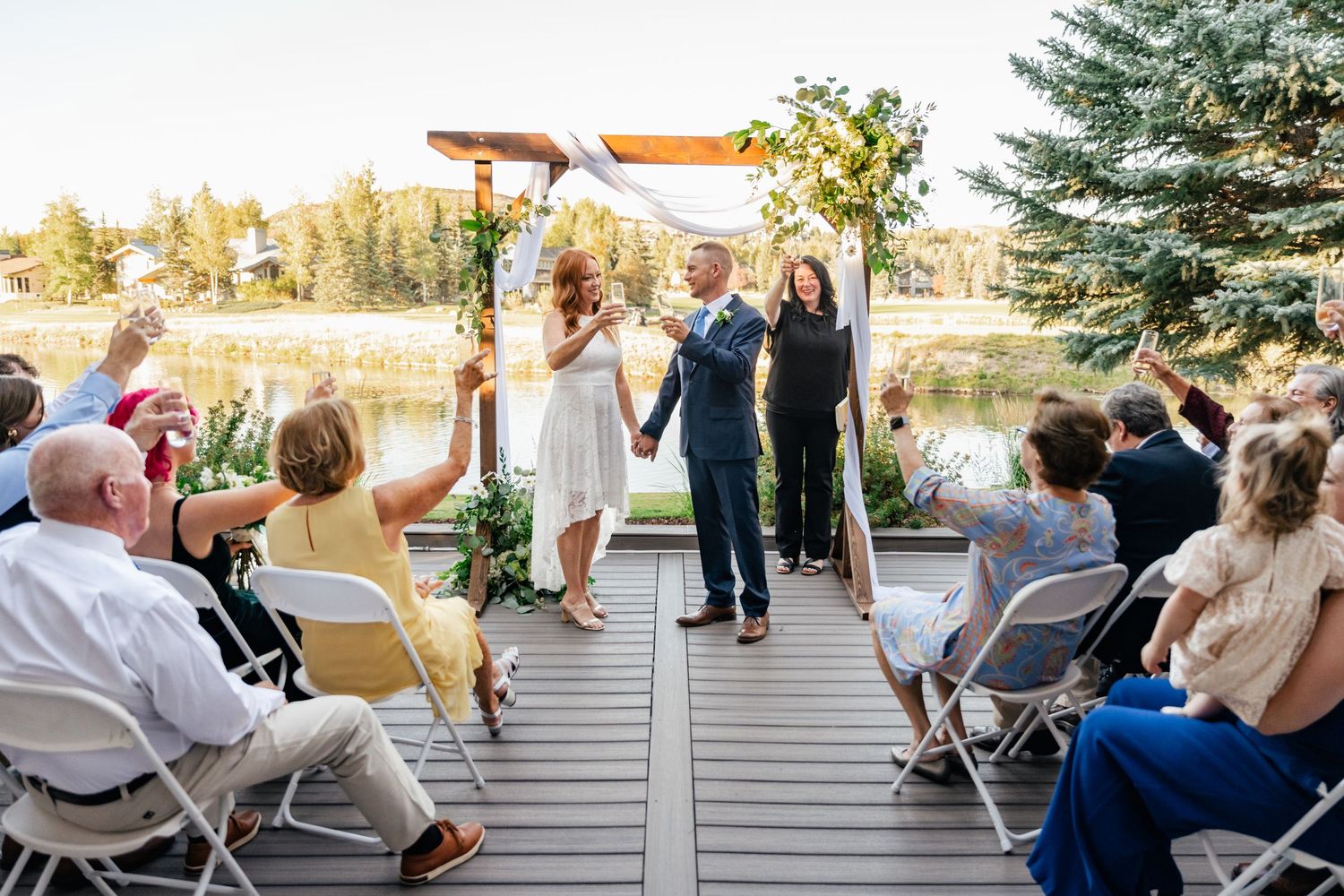 Wedding ceremony under wooden arch on deck with seated guests and pine trees in background.
