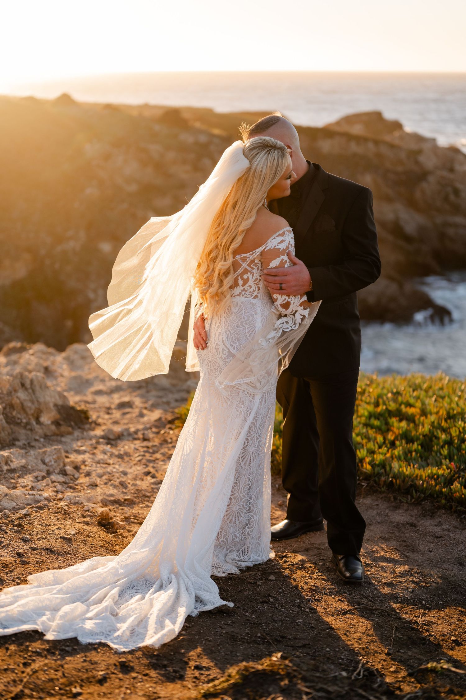 Wedding couple embraces on coastal cliffs during sunset with ocean waves and golden light creating a dreamy atmosphere.
