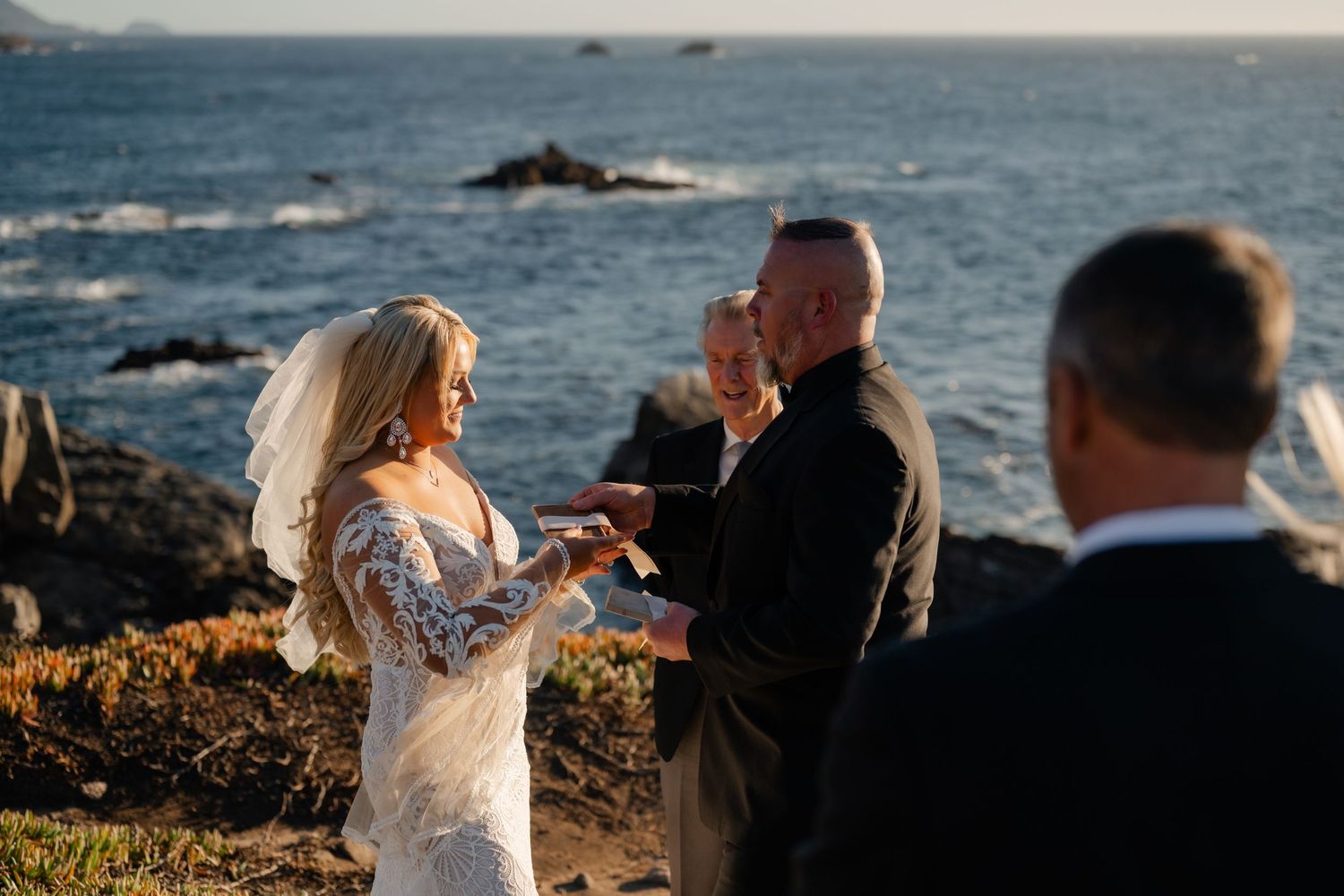 A couple exchanges rings during their wedding ceremony on a scenic oceanside cliff.