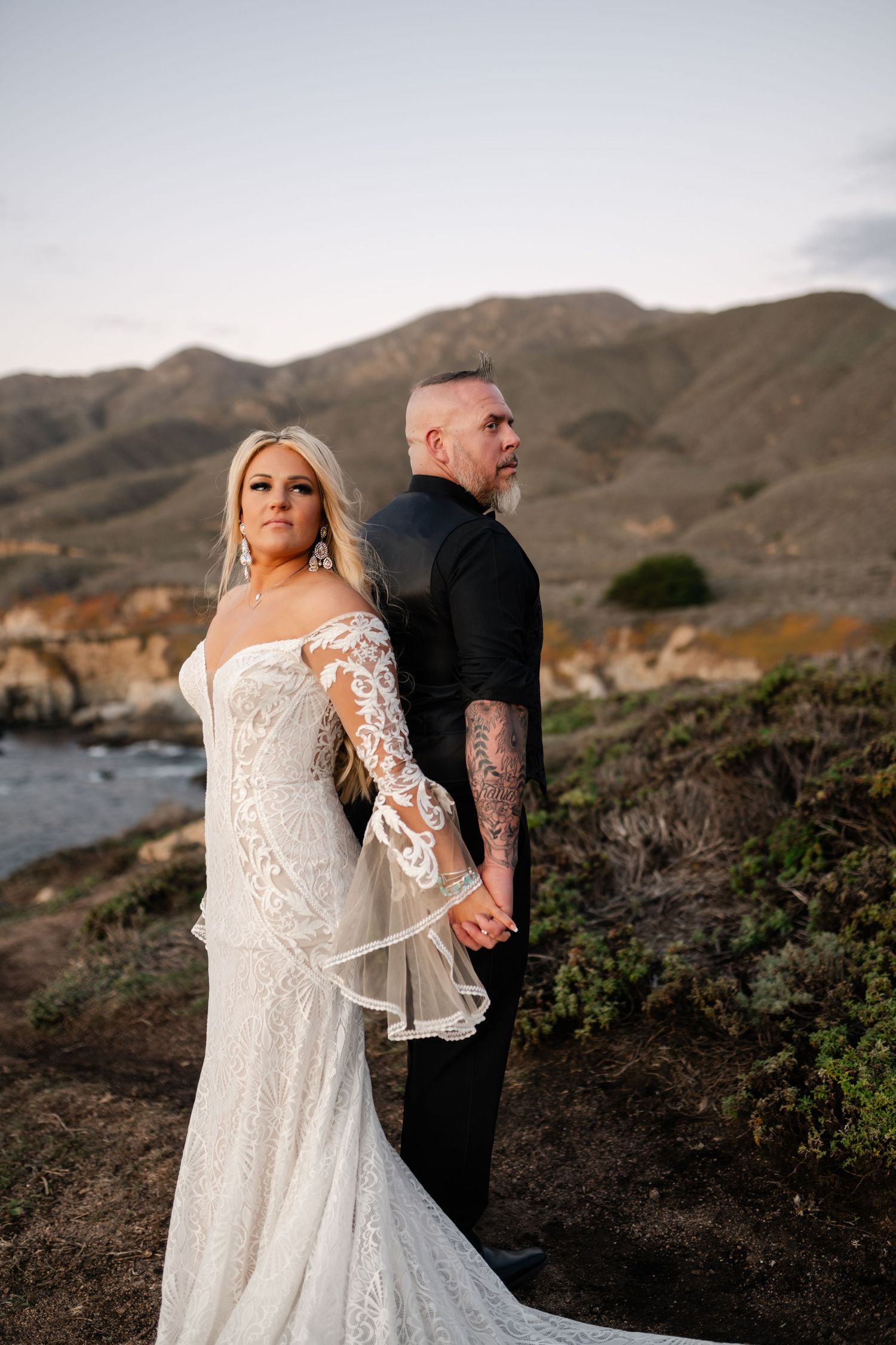 Newlyweds pose against rugged coastal landscape during sunset photo session.