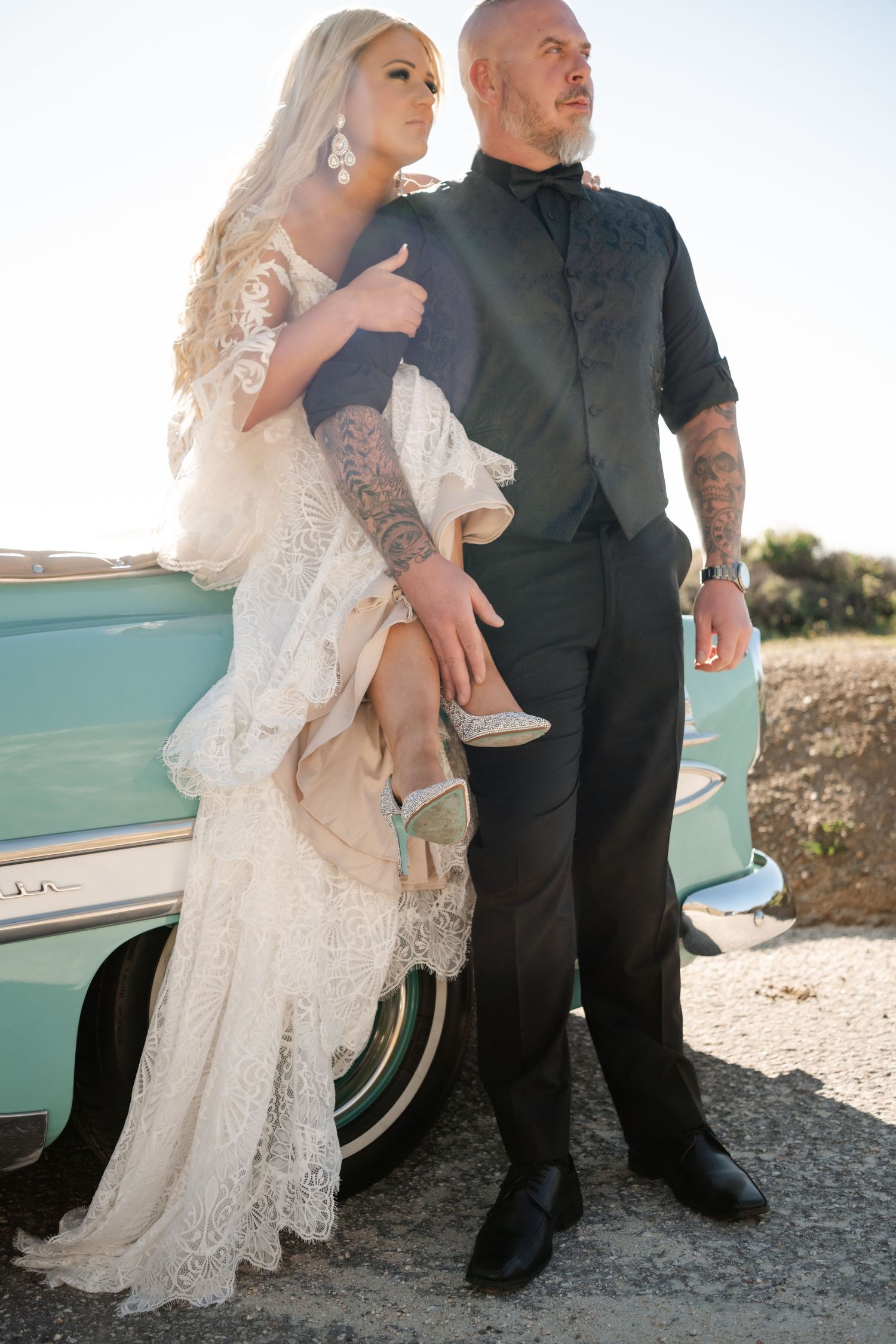A couple in wedding attire poses against a mint green vintage car during an outdoor photoshoot.
