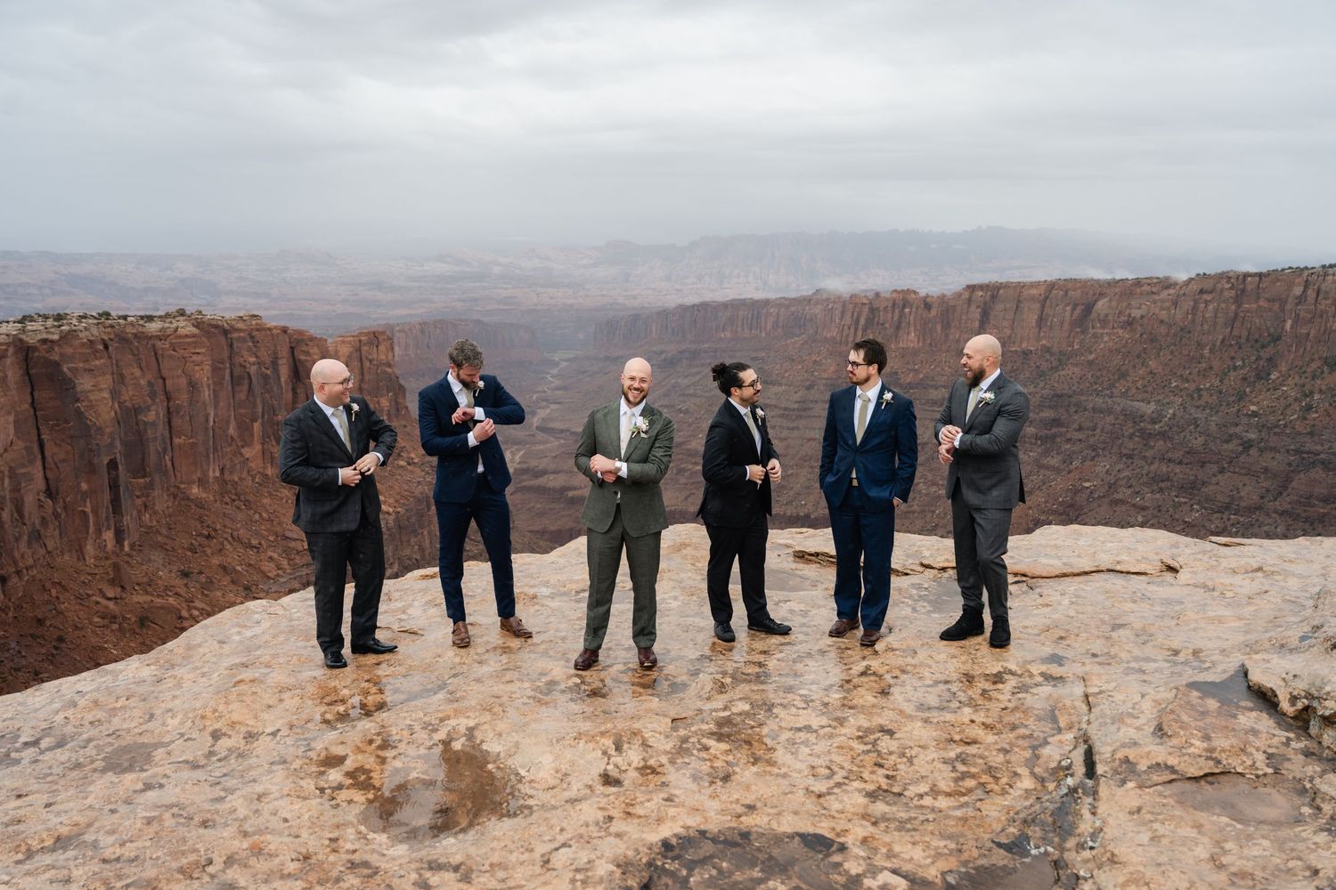 A wedding party stands at the edge of a dramatic canyon overlook with red rock cliffs stretching into the distance.