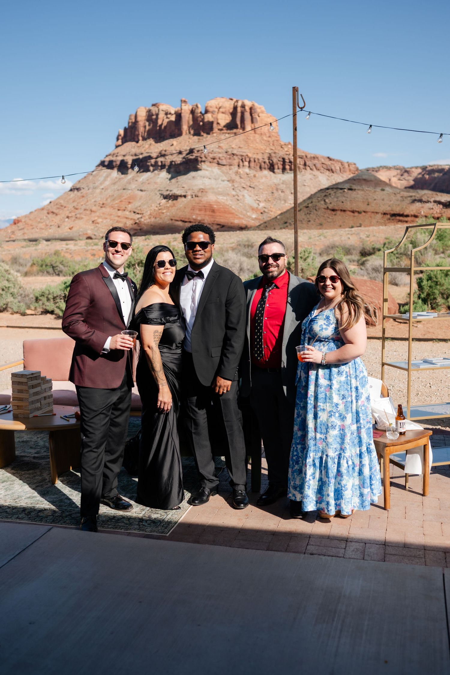 Five elegantly dressed guests pose together at an outdoor desert wedding venue with red rock formations in the background.