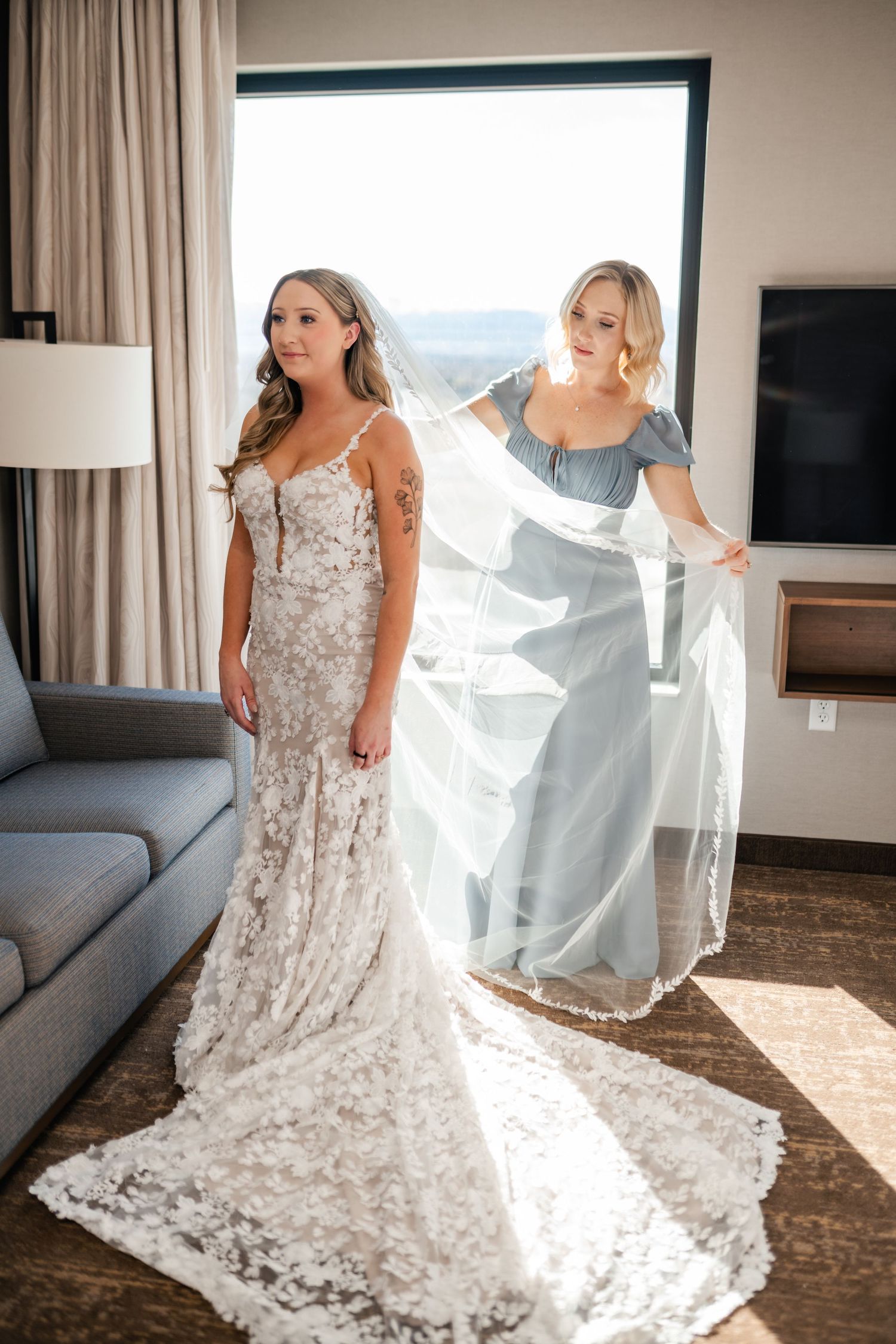 A bride in a lace wedding gown with flowing train stands while an attendant in blue adjusts her veil in sunlit room.