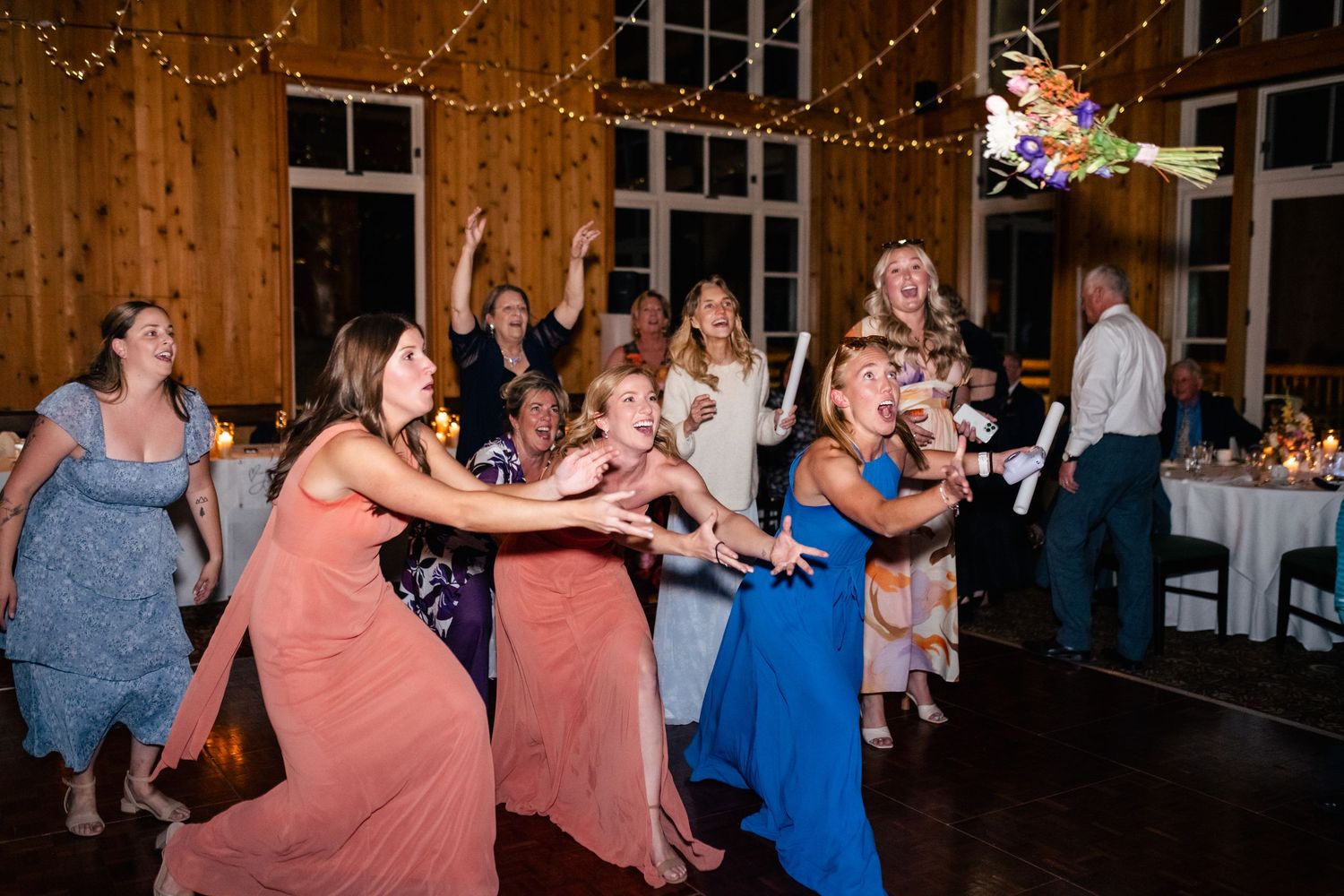 Wedding guests in colorful dresses excitedly dance and reach for a bouquet toss under string lights at a rustic reception venue.
