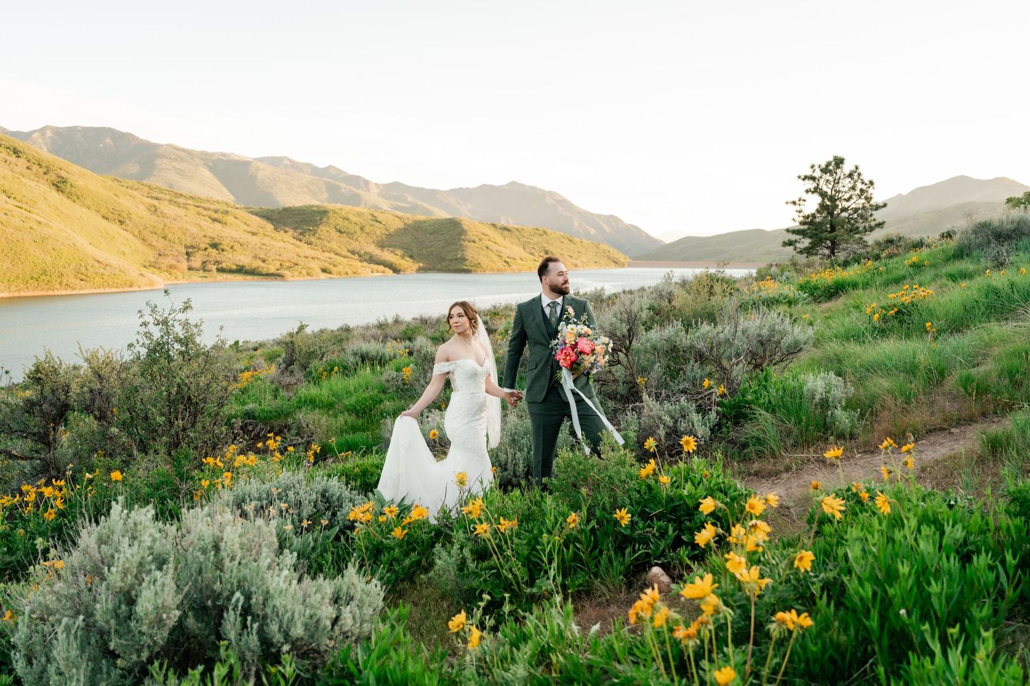 Wedding couple walks through mountain wildflower meadow with lake view in background.