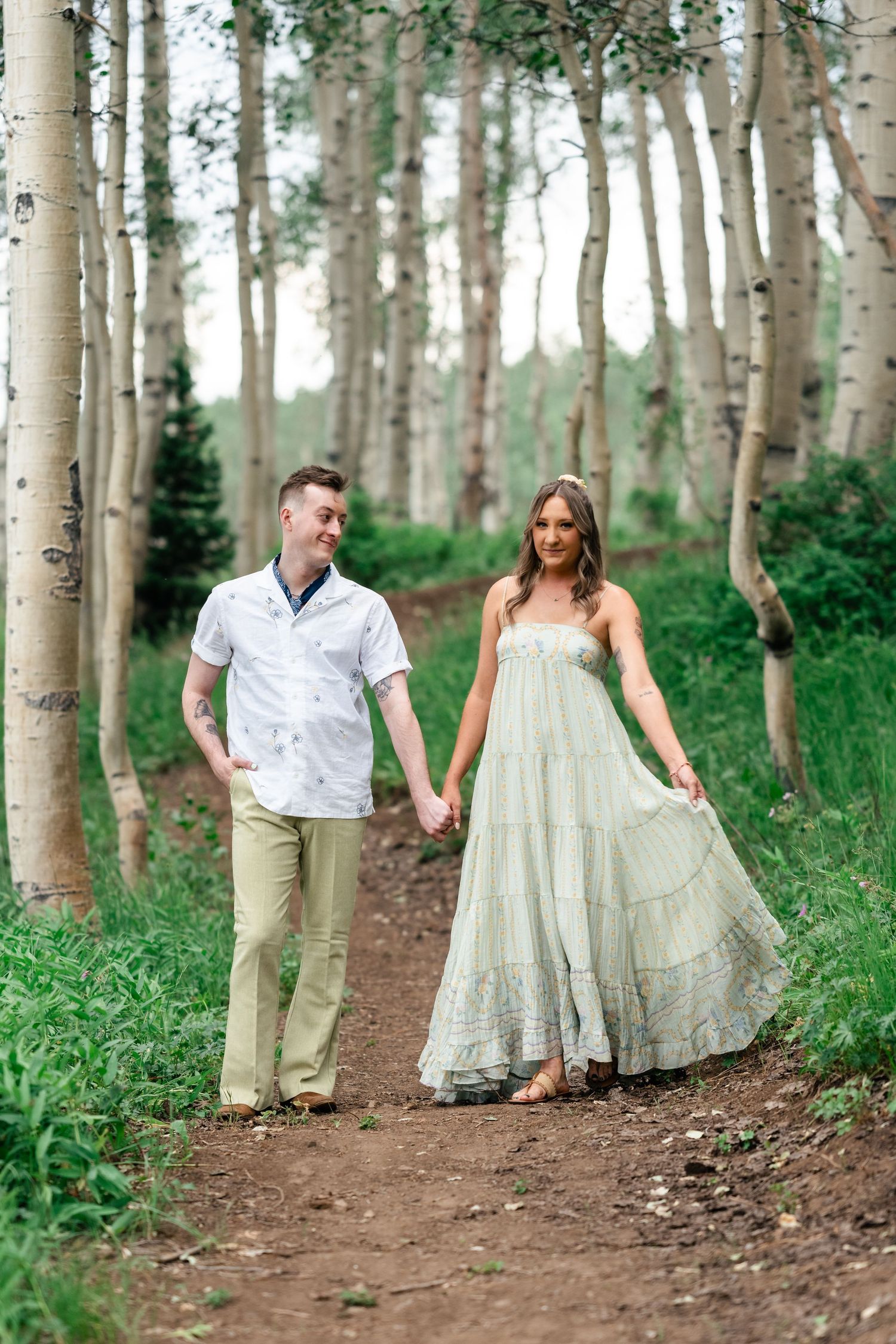 A couple walks hand in hand along a dirt path through a scenic aspen grove on a sunny summer day. Utah mountain engagement session