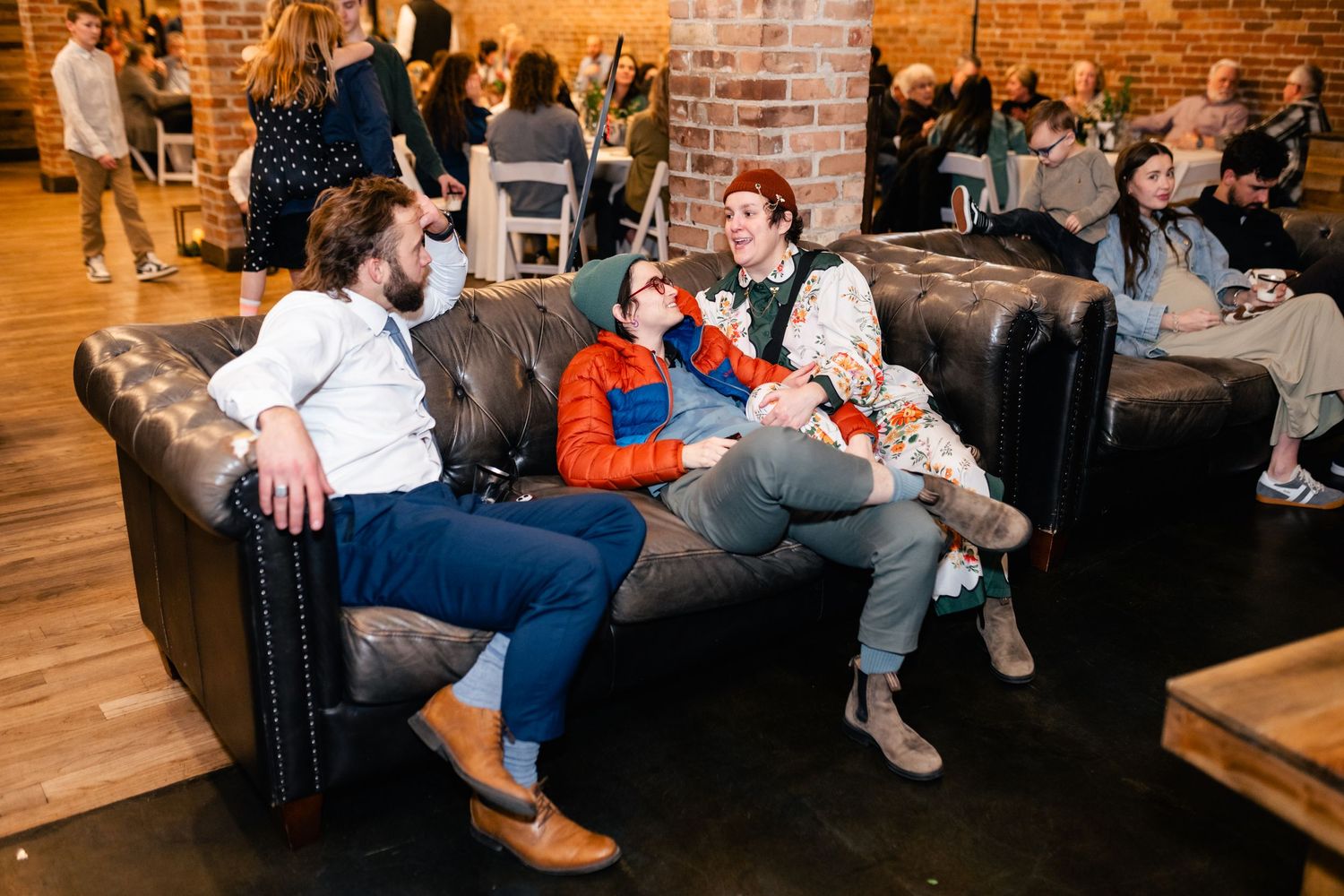 People relax on a brown leather couch in an industrial-style room with exposed brick walls and hardwood floors.