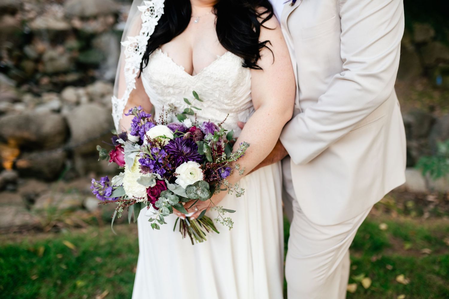 A bride in a white dress holds a stunning purple and white bouquet while posing with her groom.