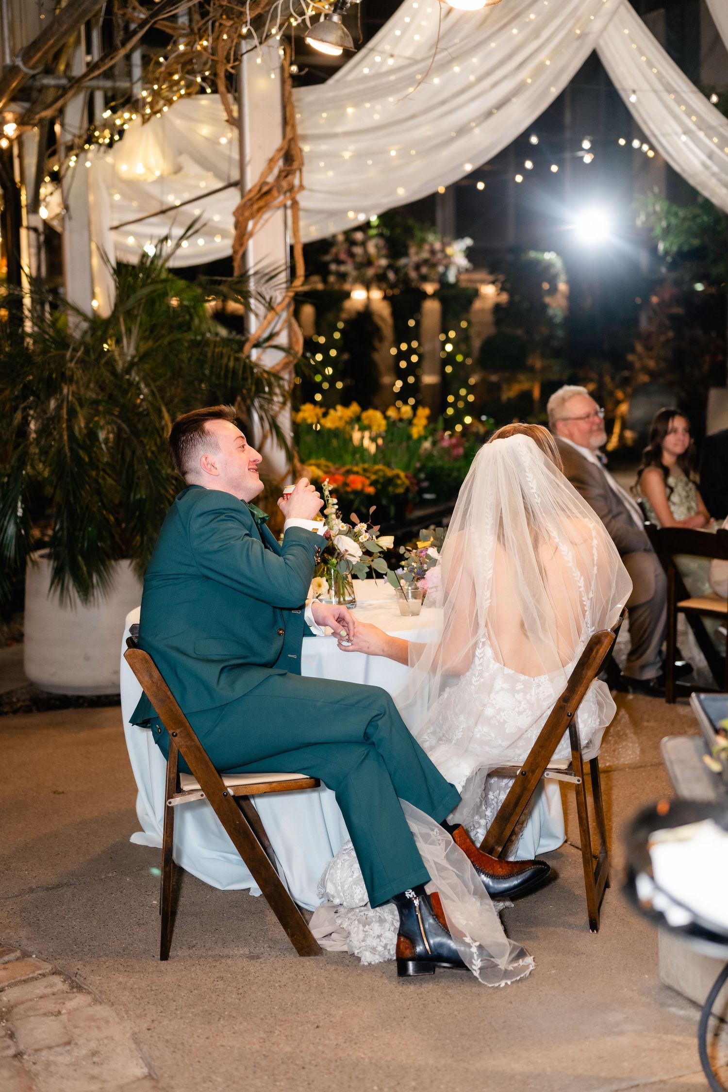 Wedding guests enjoy a reception celebration under twinkling lights and draped fabric in an evening outdoor venue setting.