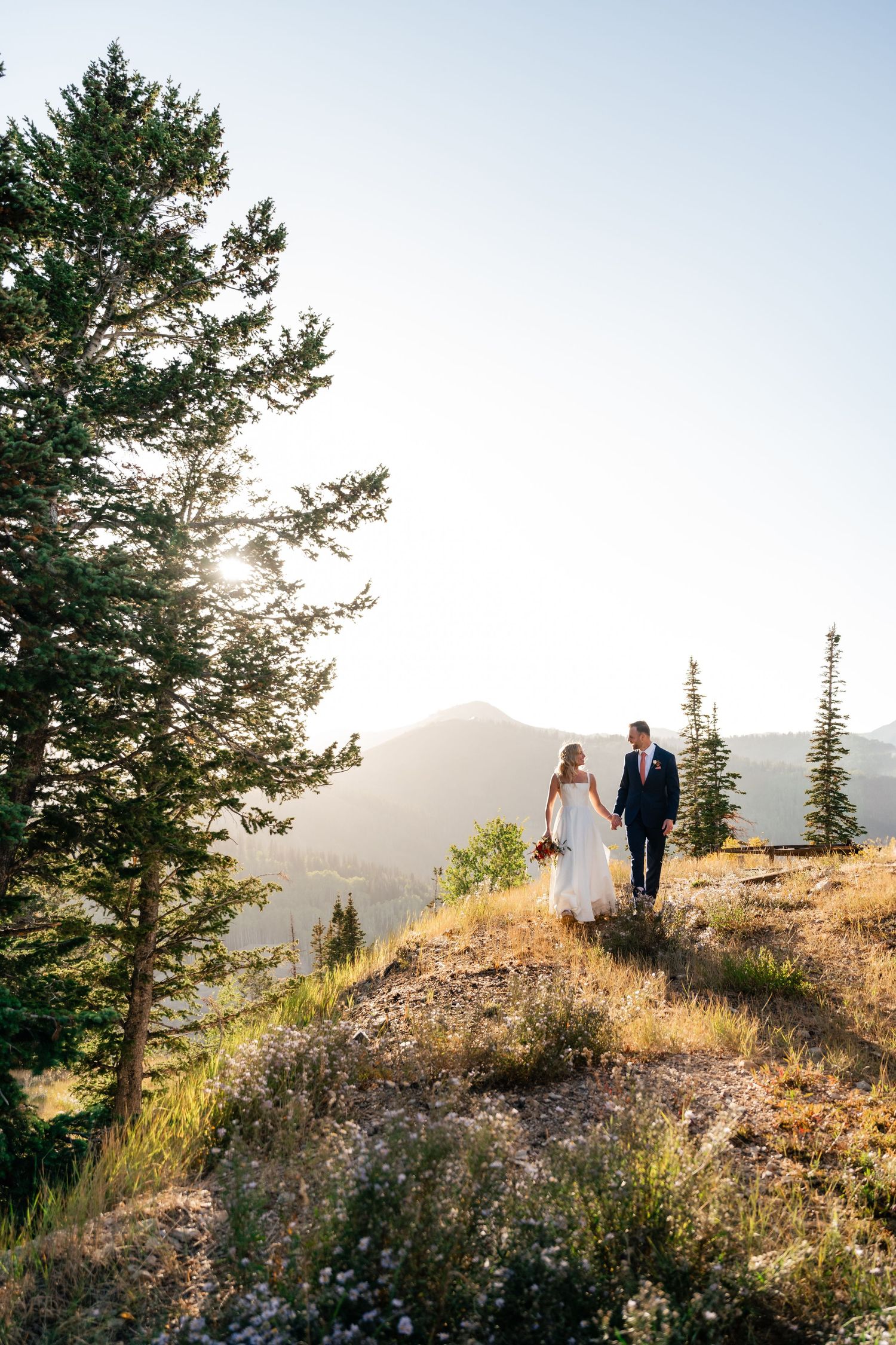 Couple walks hand in hand along mountain trail during sunset with pine trees in background.