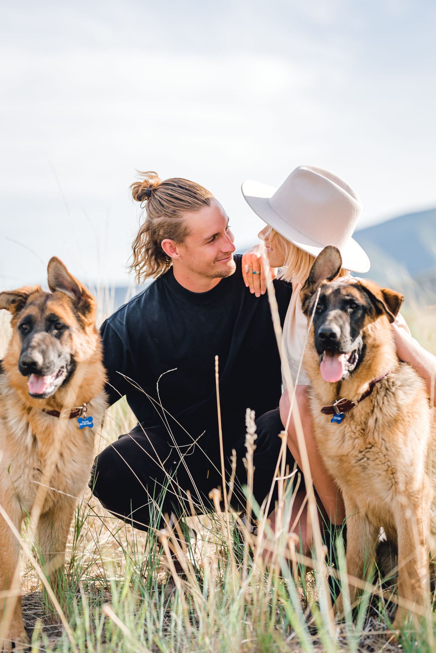 Couple with two German Shepherd dogs enjoying a tender moment in tall grass outdoors.