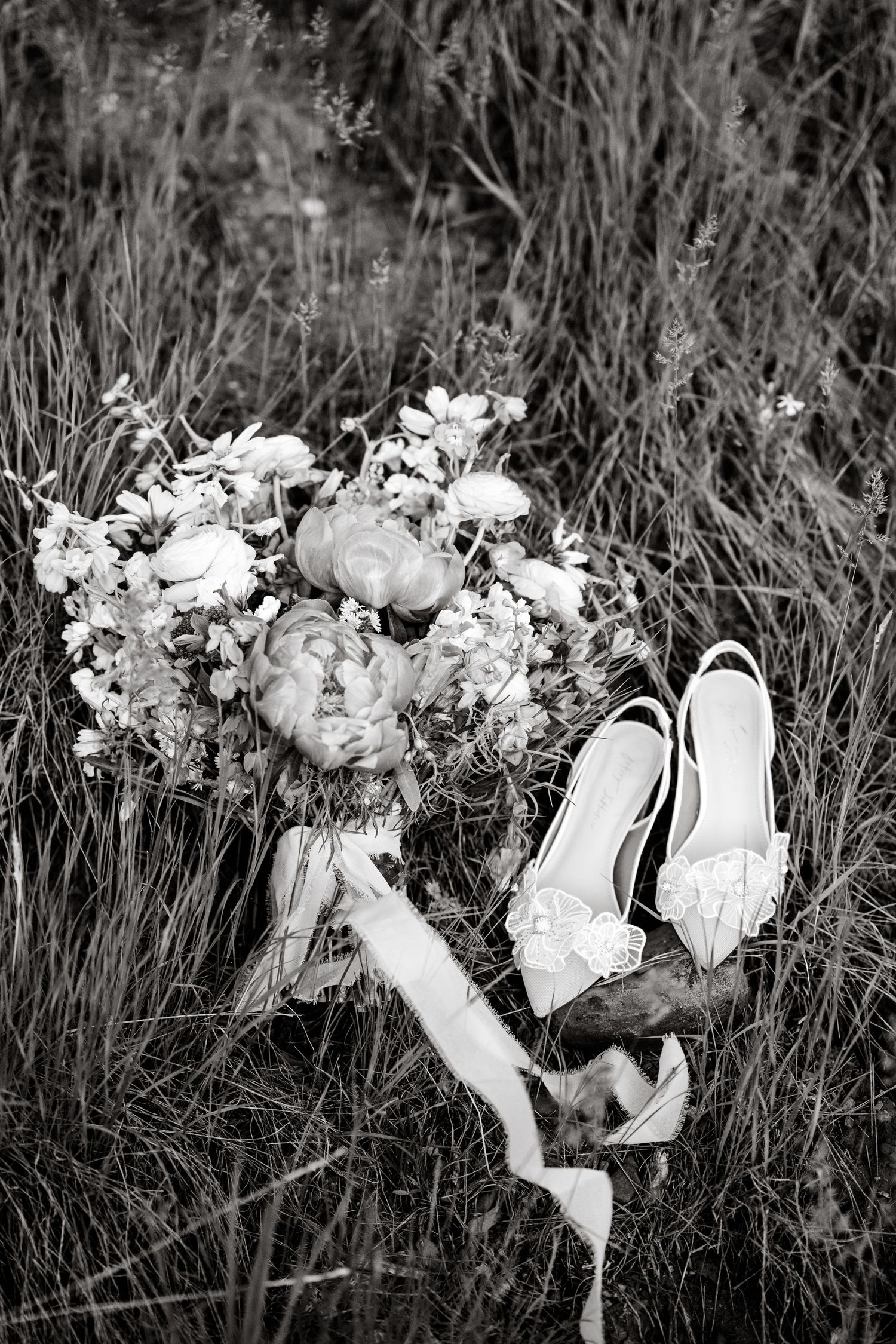 A black and white detail shot of elegant wedding shoes surrounded by delicate floral arrangements.