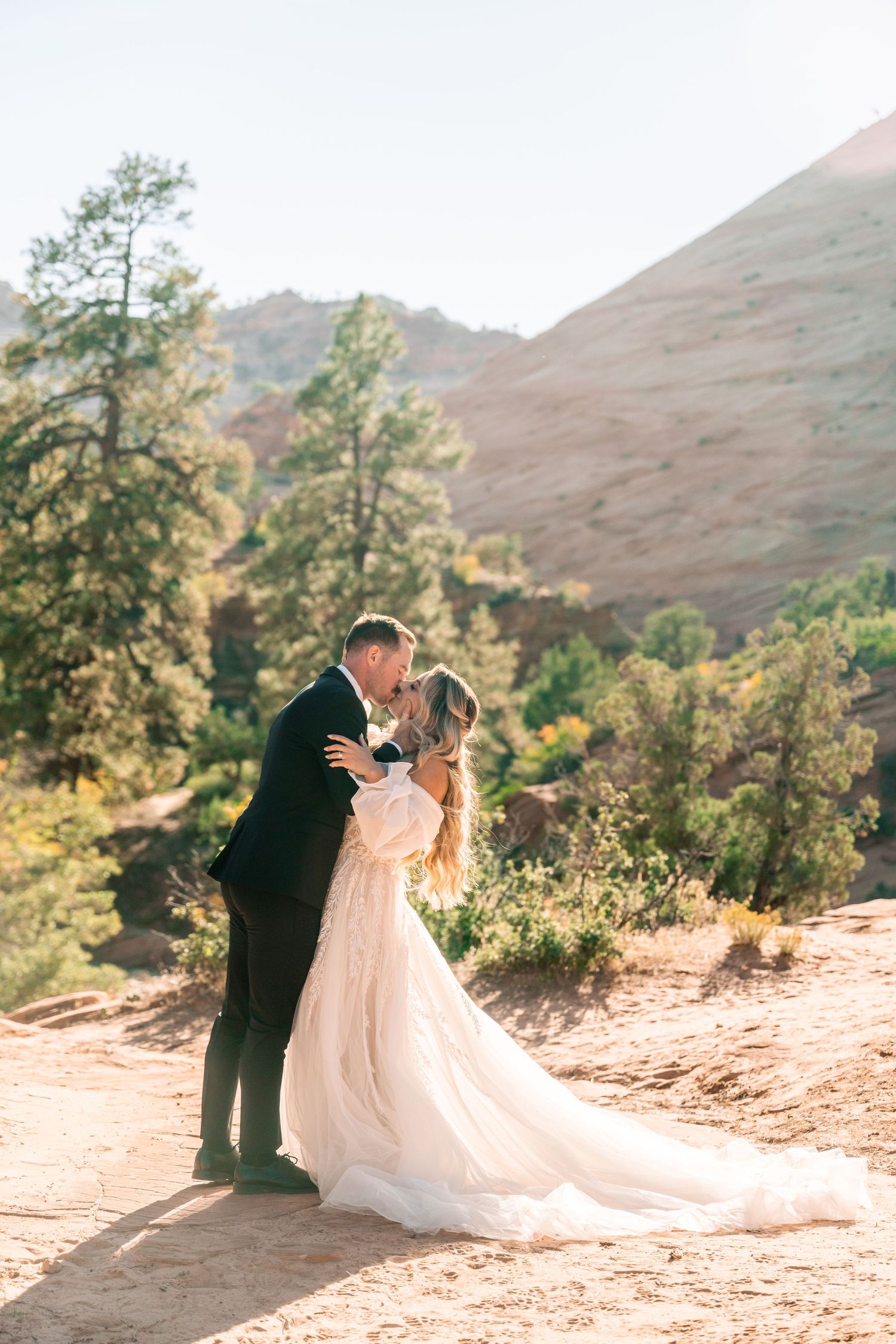 A couple shares a romantic moment on a rocky trail surrounded by mountains and pine trees.