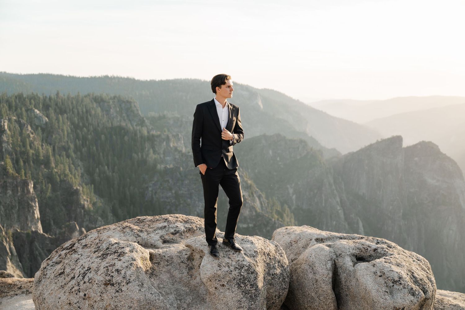 A man in a black suit stands on a rocky summit with sweeping mountain valley views at golden hour.