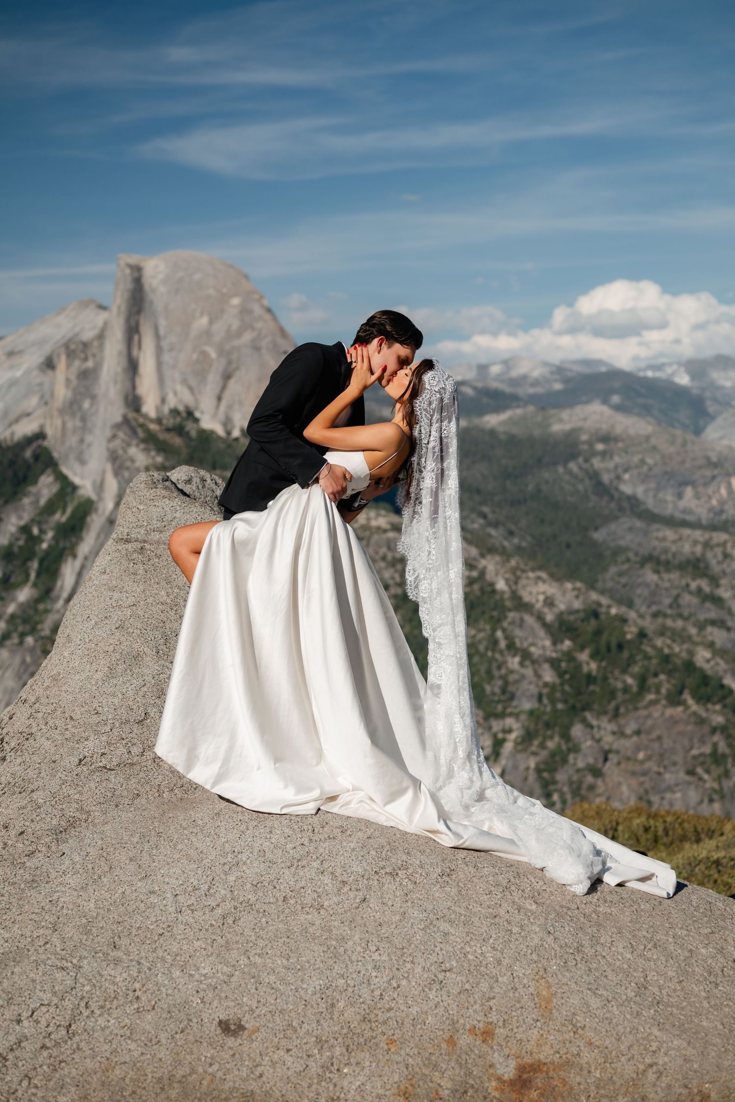 Bride and groom share a romantic kiss on a rocky overlook with Half Dome and Yosemite mountains in the background.
