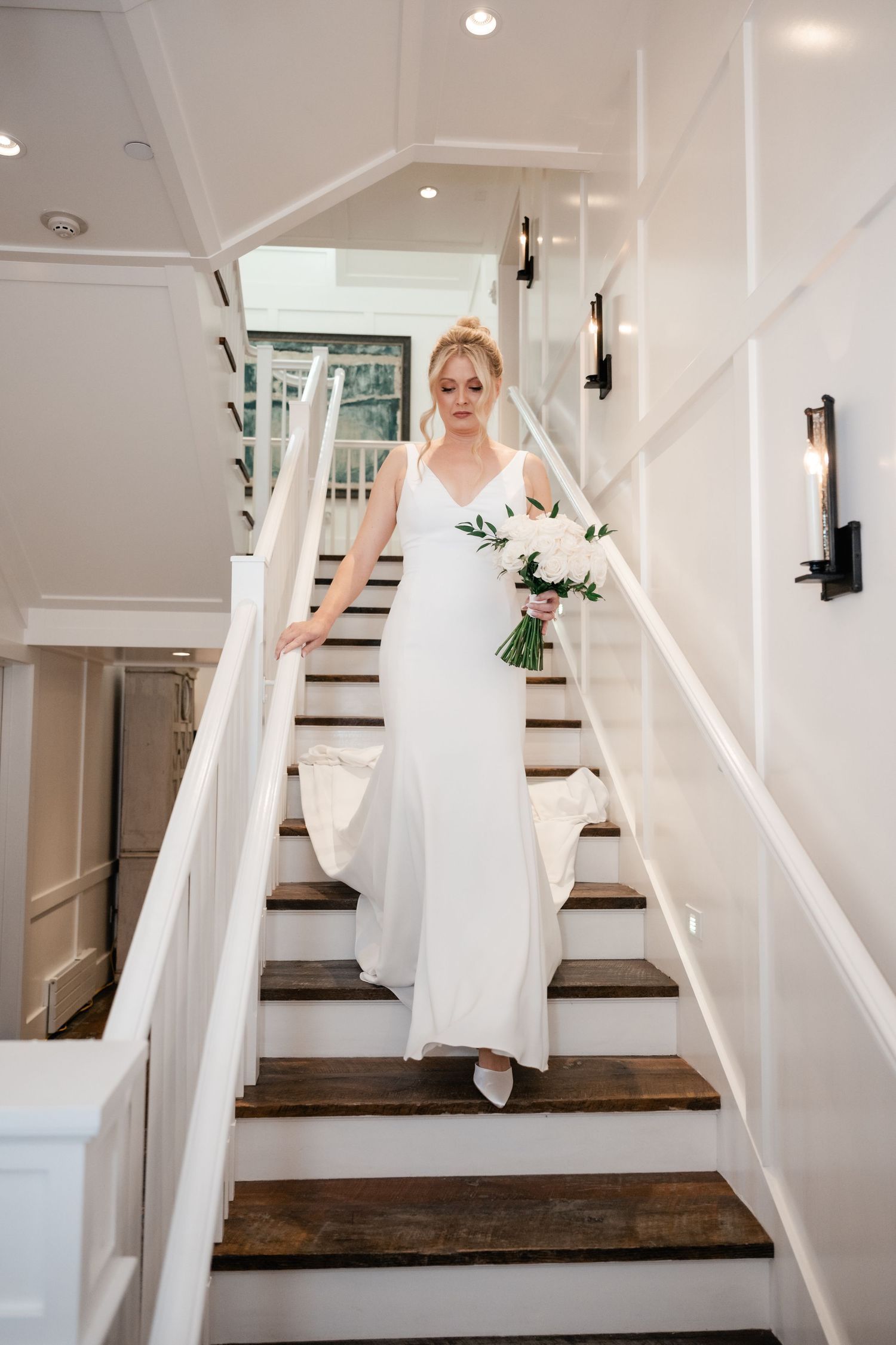 Bride in white gown descending wooden staircase holding white floral bouquet in elegant bright interior of the Washington School House Hotel