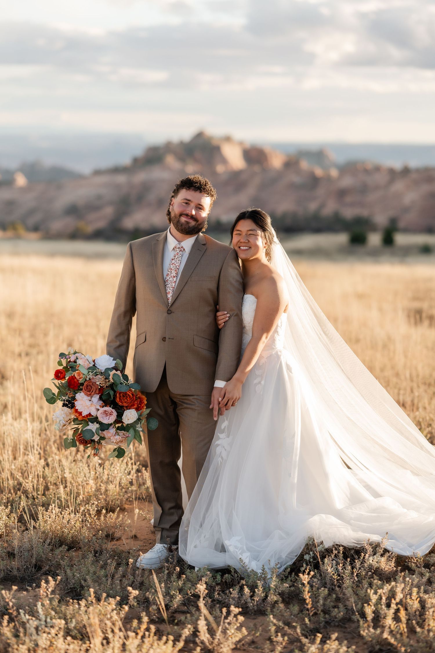 A couple poses in a field at sunset, with the bride's white dress flowing dramatically in the breeze.
