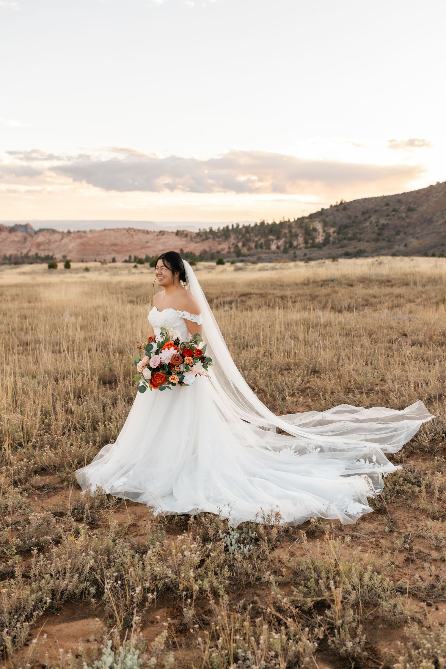 Bride in flowing white gown poses dramatically in golden mountain field with red floral bouquet.