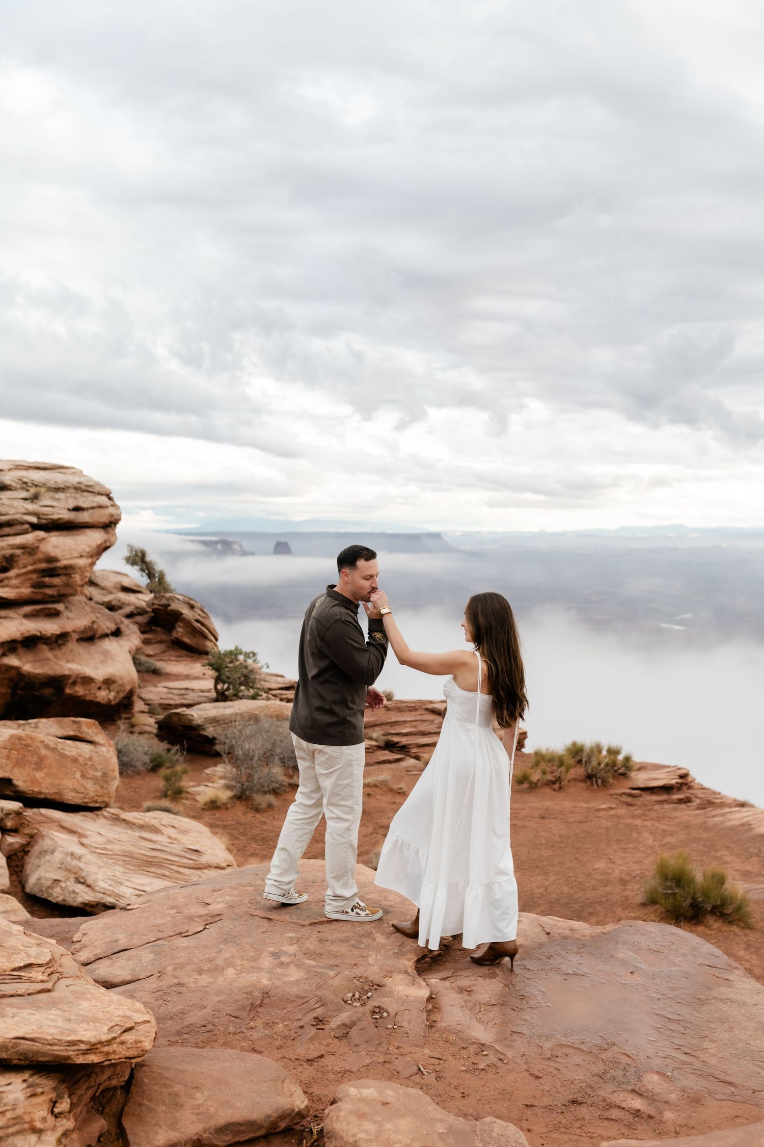 A couple dances together on a scenic red rock overlook with cloudy skies in the background.