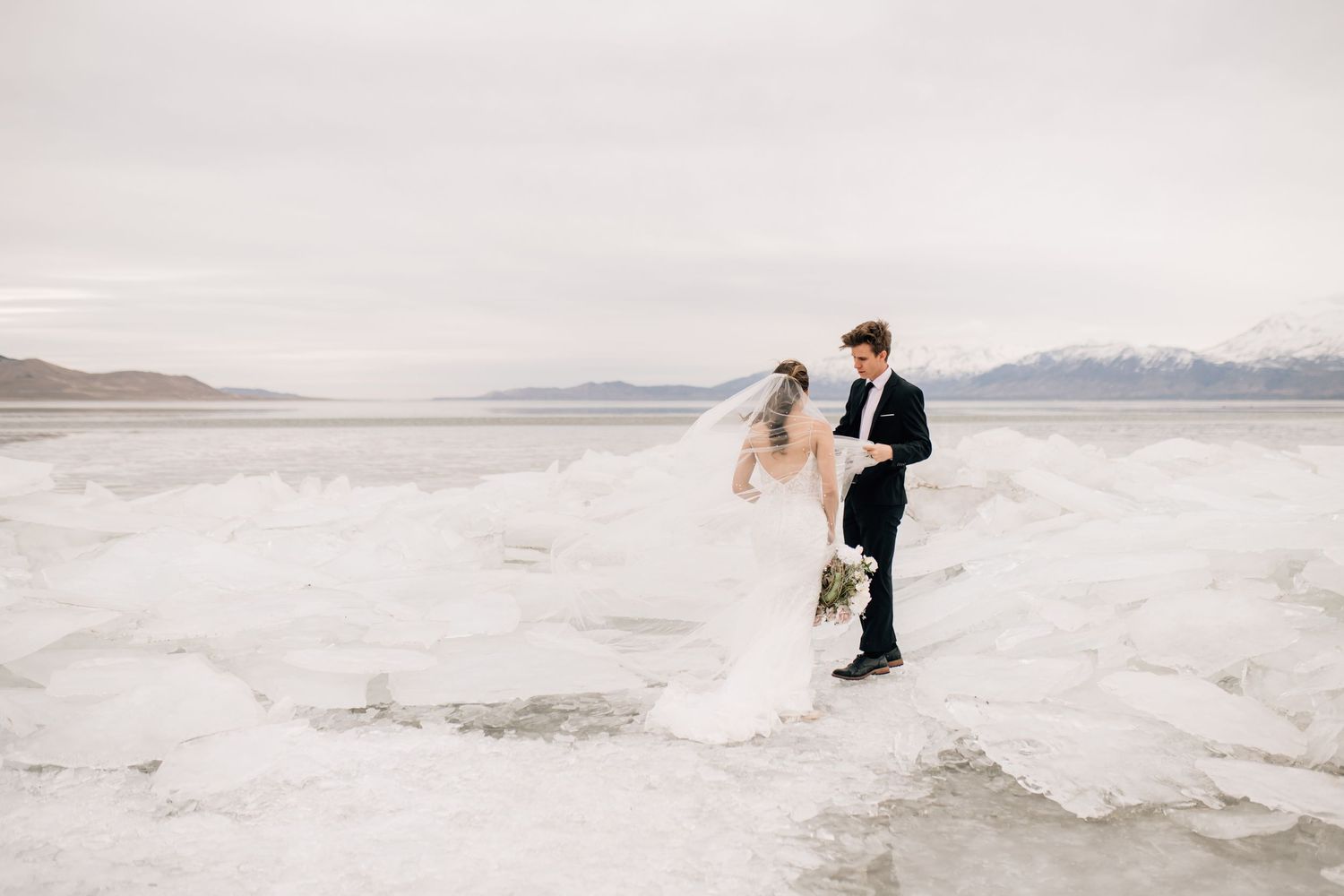 A couple in wedding attire walks together on a snowy white beach landscape during a winter photoshoot. Best Utah Wedding Photographers.