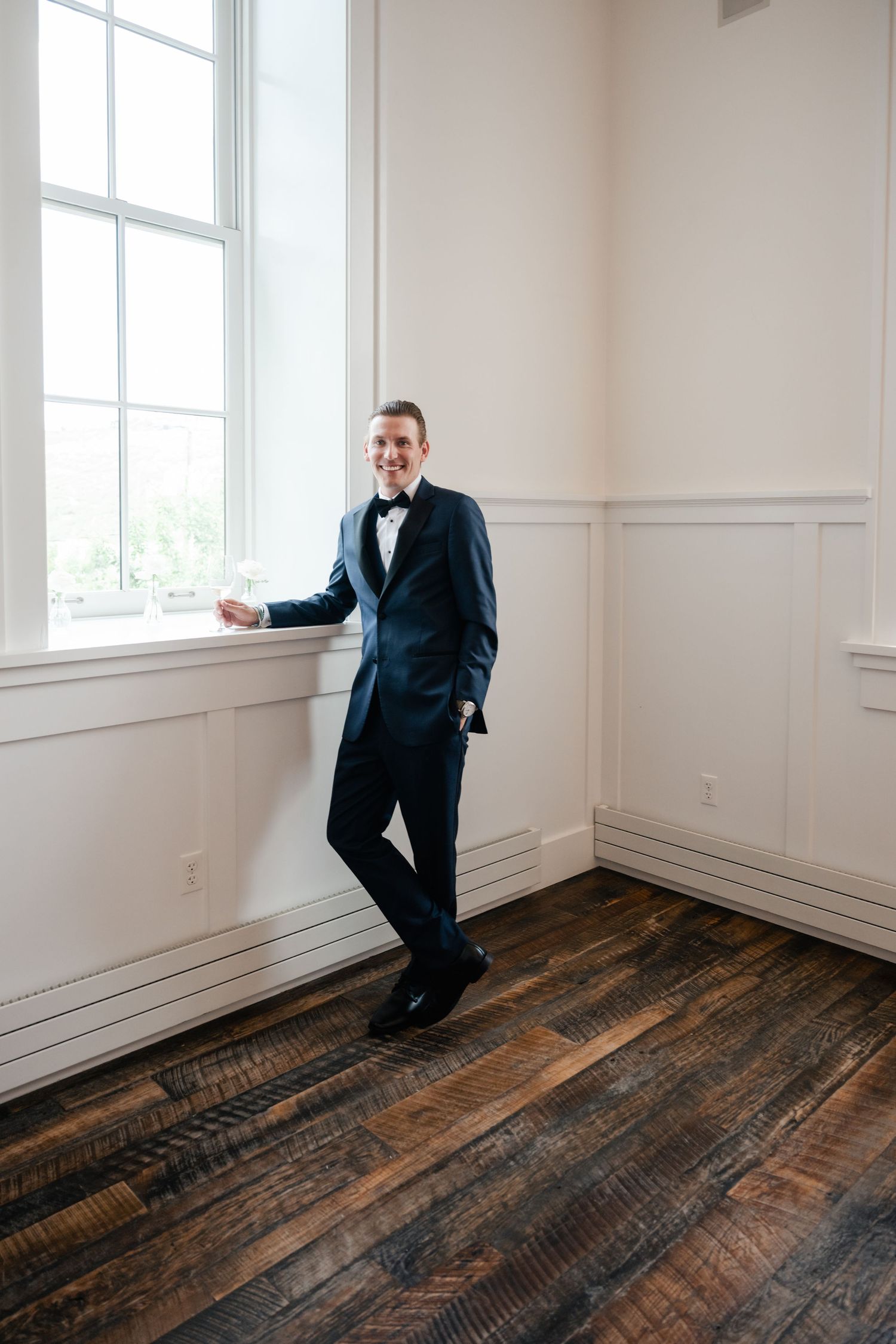 Groom in black tuxedo poses playfully in minimalist white room with hardwood floors.