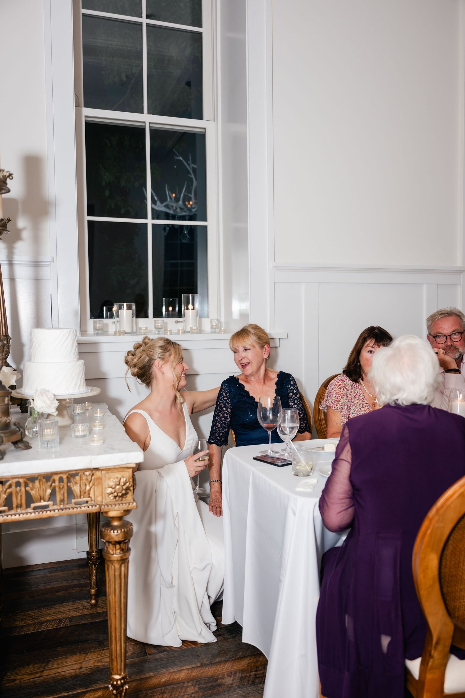 Wedding guests mingle and chat in an elegant white room with tall windows and classic decor.