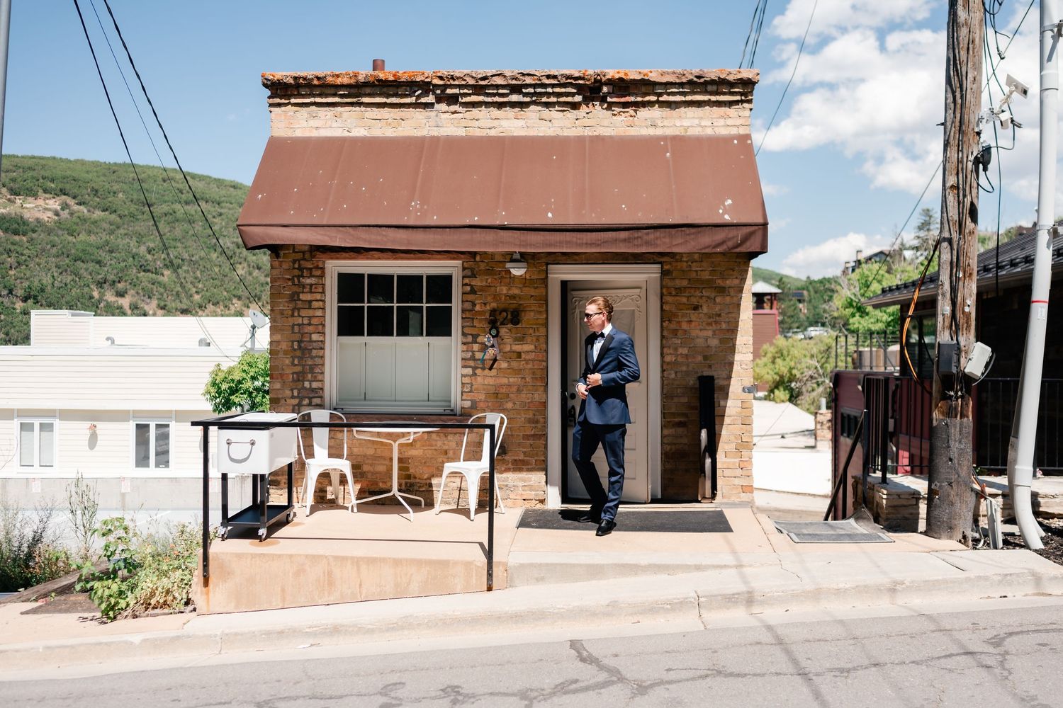 Small brick storefront with brown awning and porch in a mountain town setting.