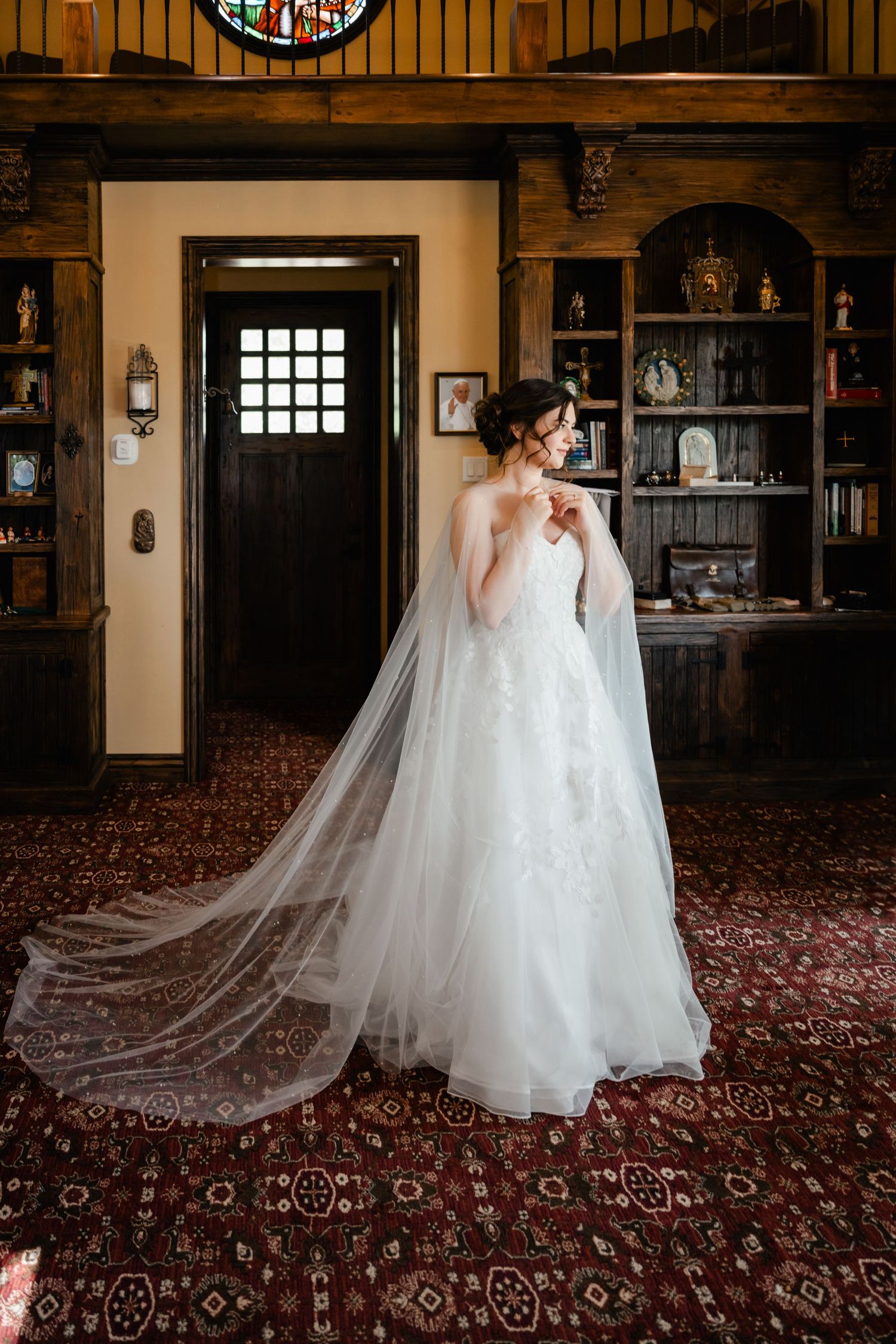 Bride in flowing white gown and veil stands in elegant room with patterned carpet.