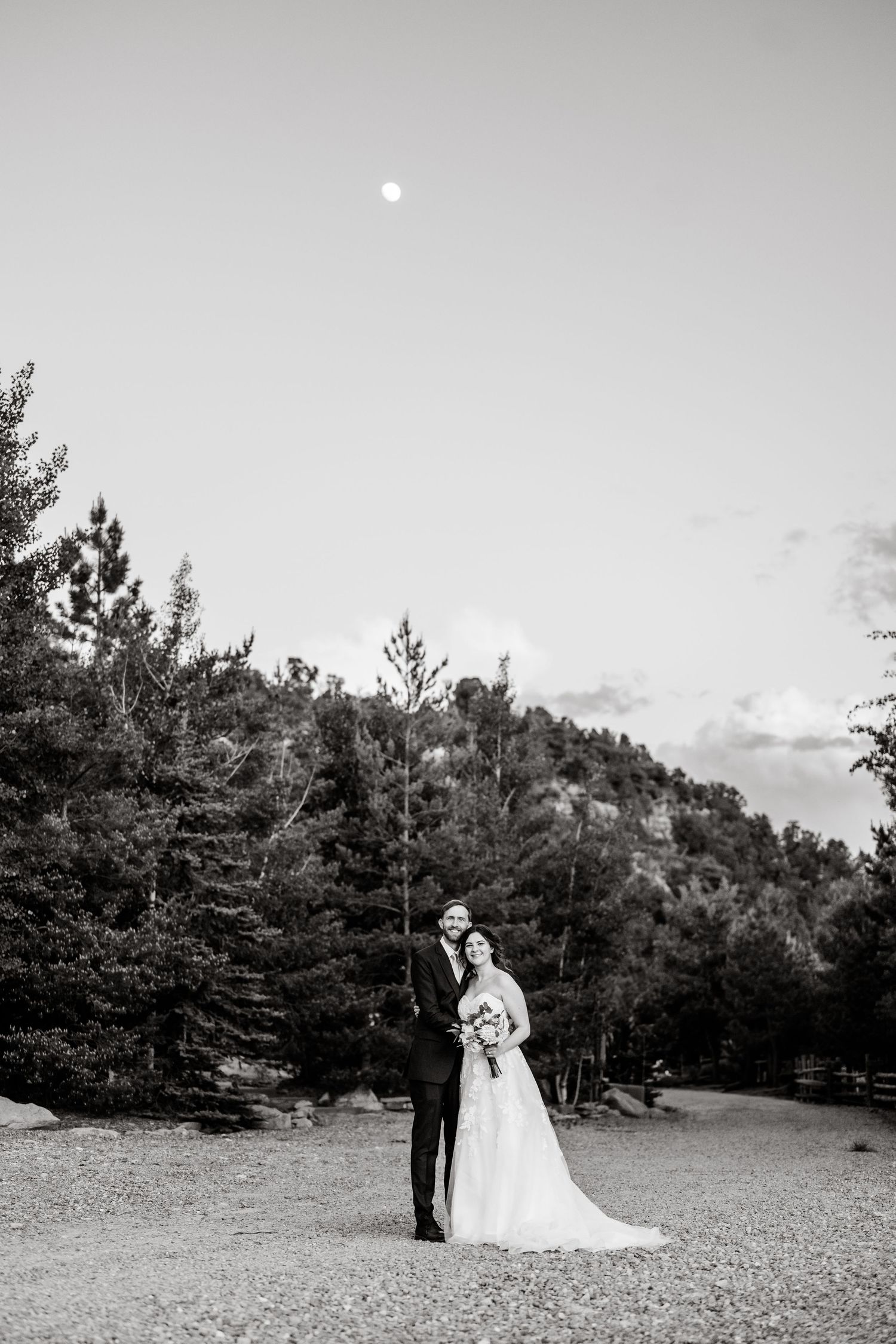 A couple in wedding attire stands together under a full moon in a mountainous pine forest landscape in black and white.