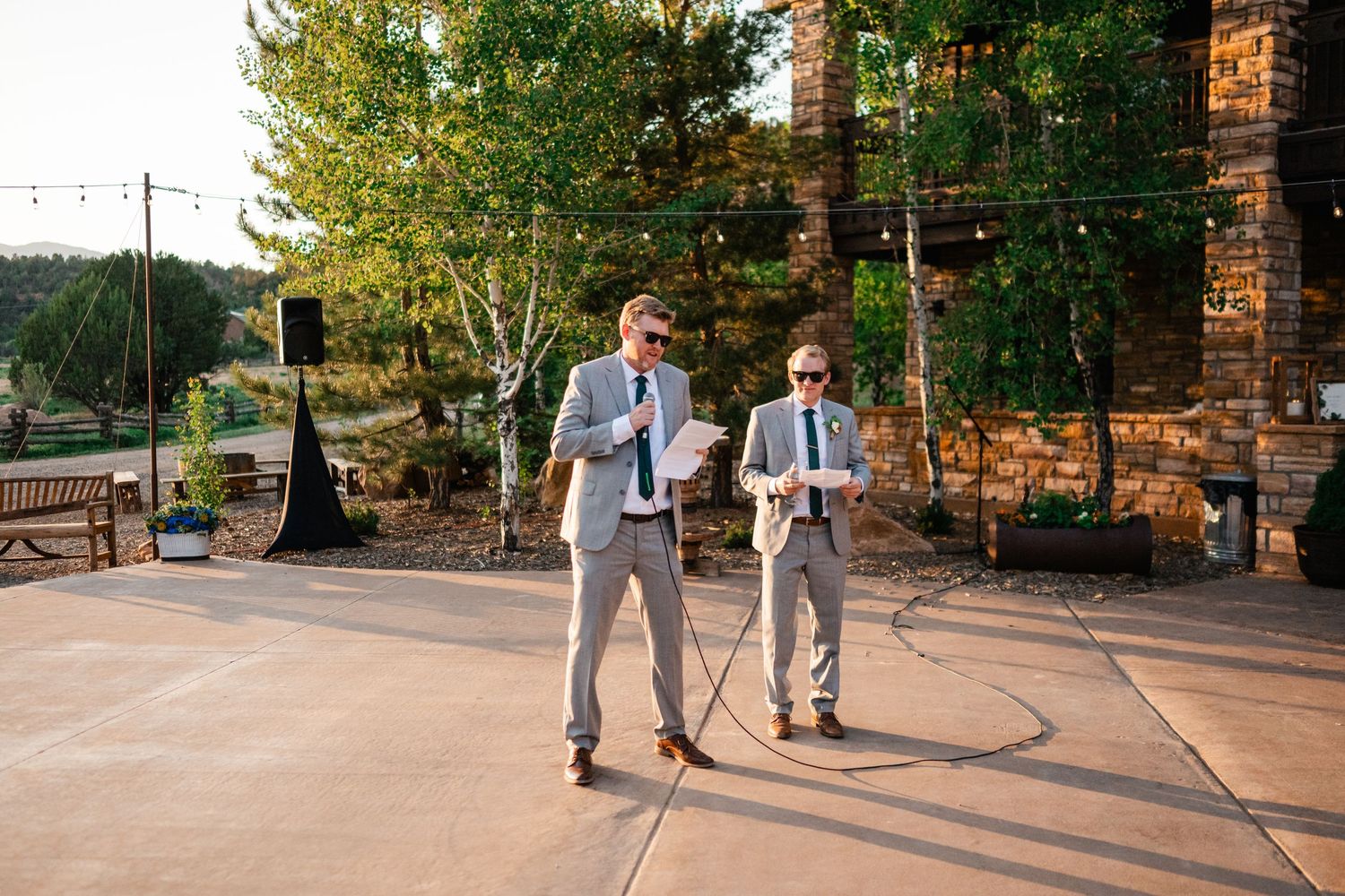 People walk and socialize on a paved outdoor path surrounded by green trees and brick buildings.