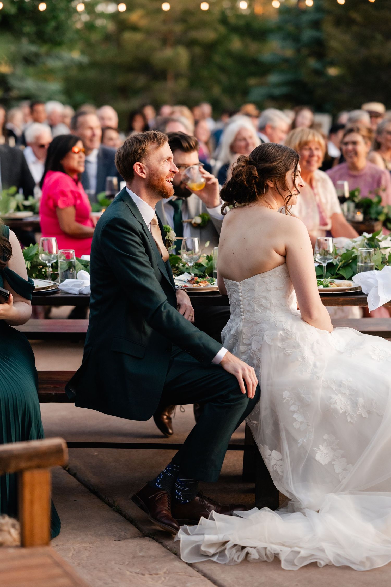Wedding couple sits on outdoor benches during reception with guests visible in background.