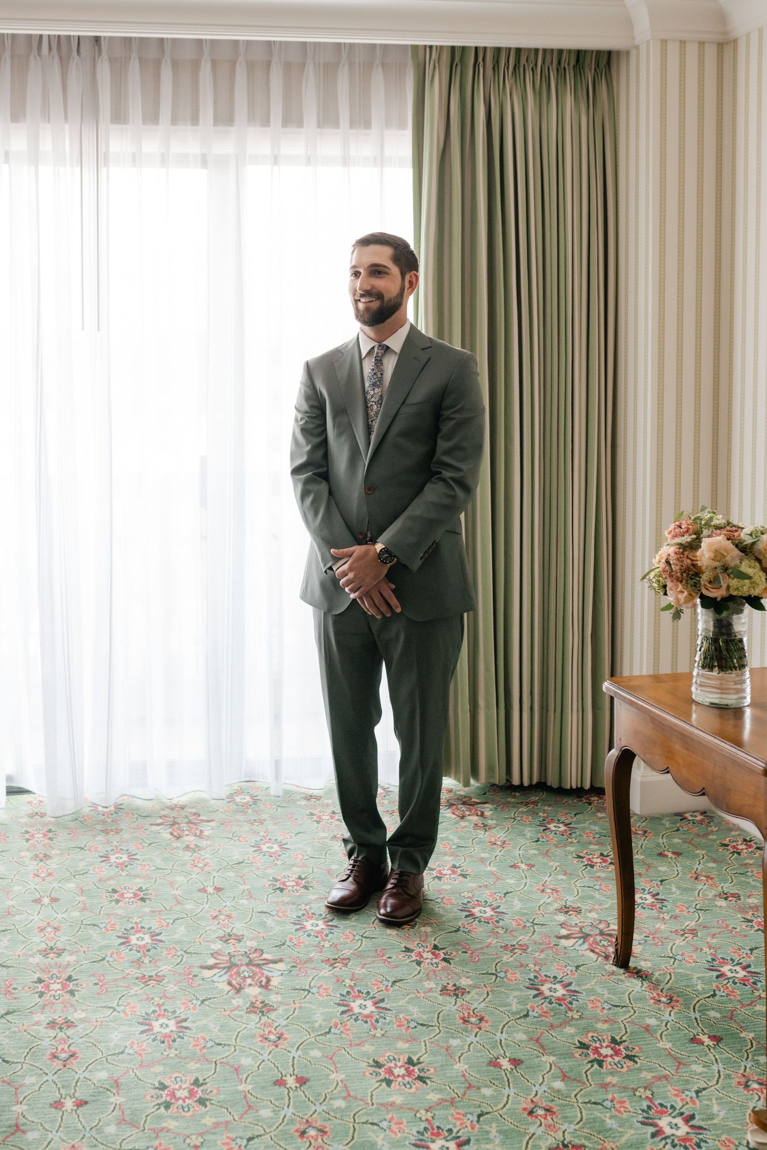 A groom in a gray suit stands in a hotel room with floral carpeting and green curtains.
