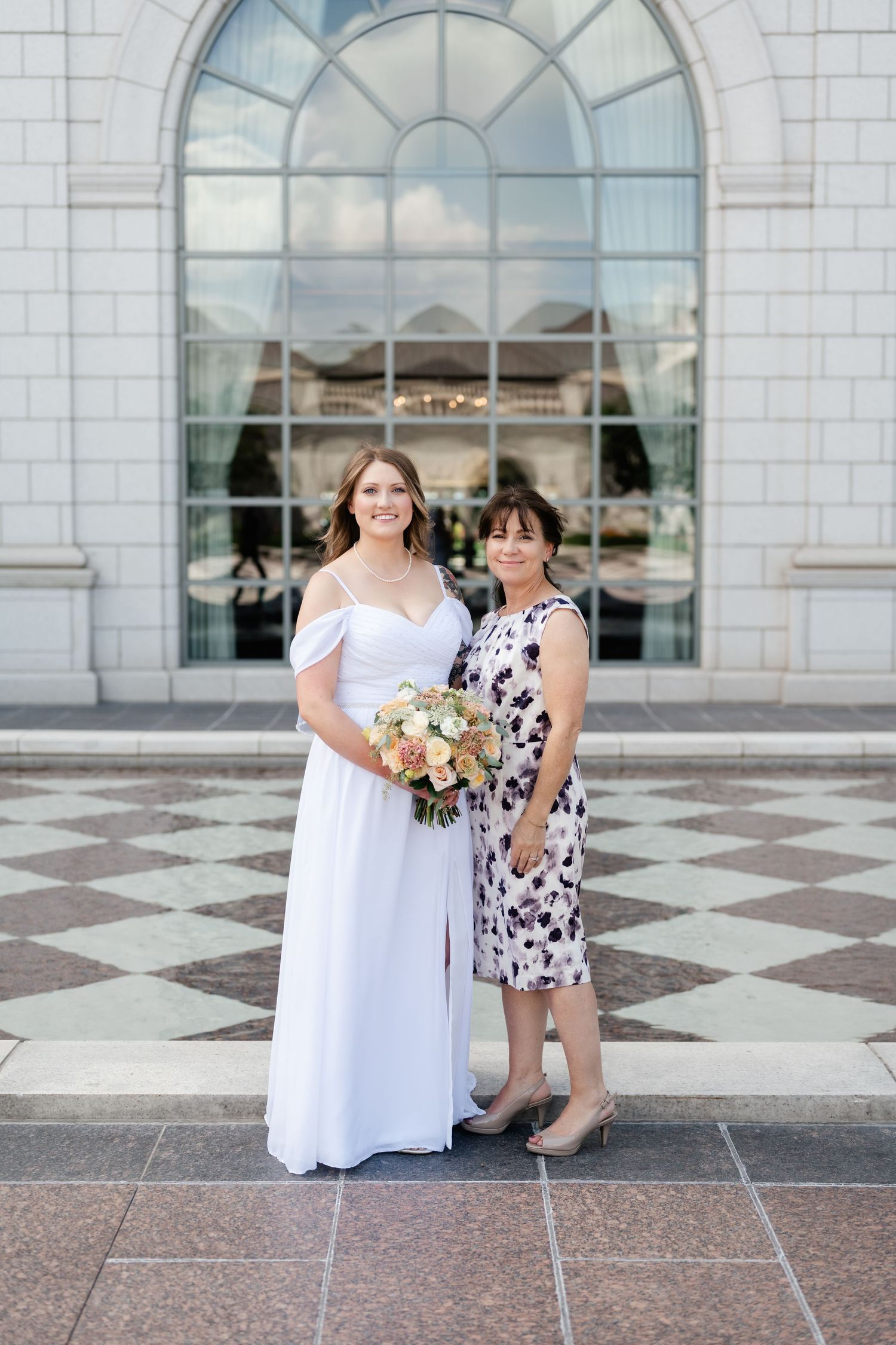 A bride in a white dress stands with a woman in a patterned dress in front of an arched window at a wedding venue.
