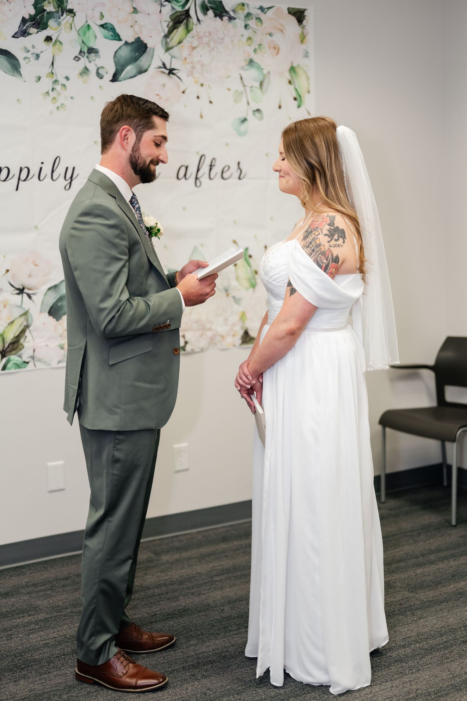 A couple in wedding attire read vows to each other in a room with floral wallpaper during their intimate ceremony.