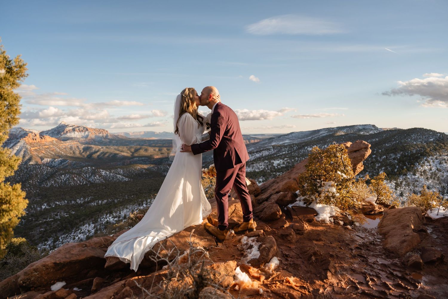 Wedding couple embracing on red rock overlook with mountain vista at sunset.