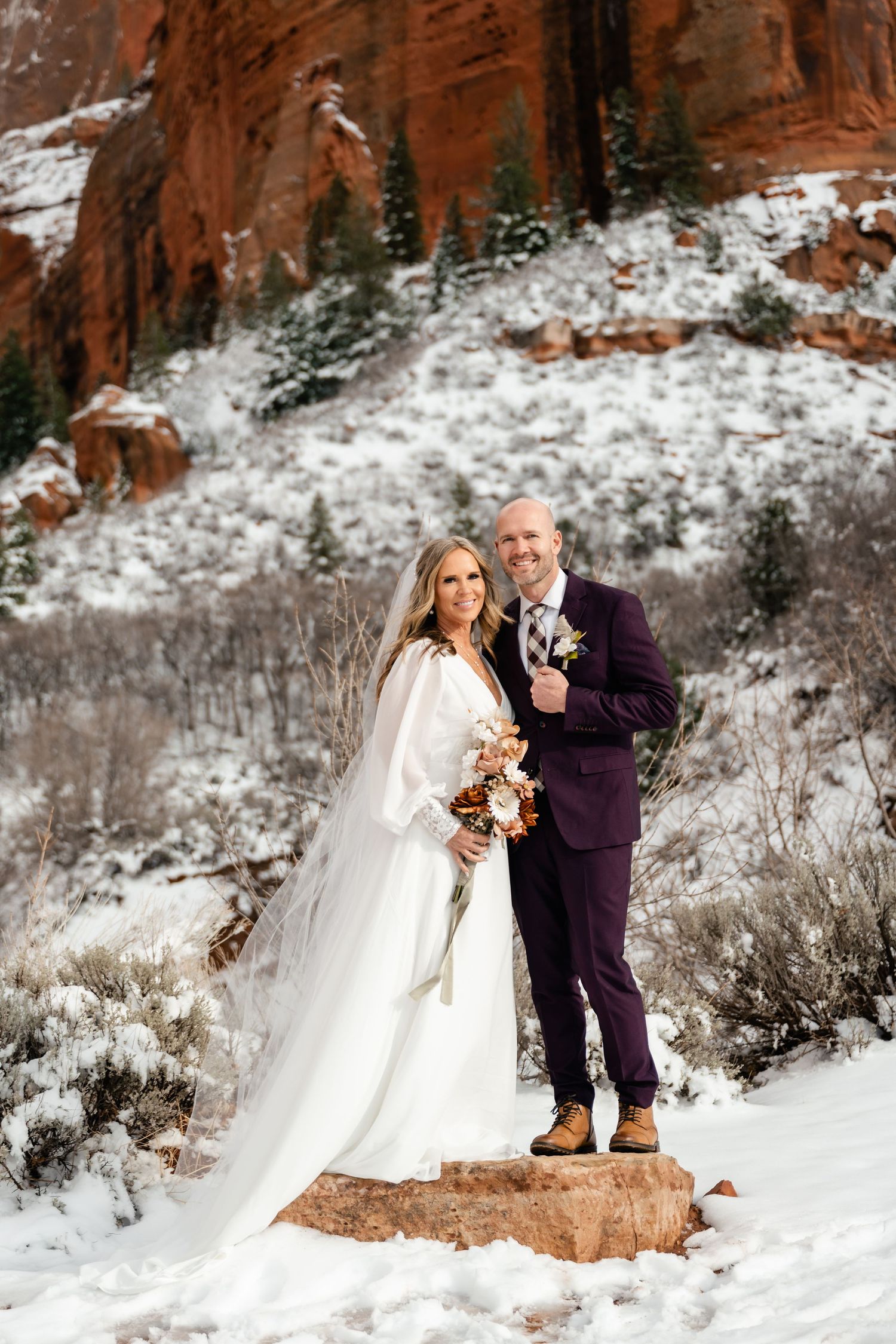 Wedding couple poses on snowy red rock formation in winter landscape.