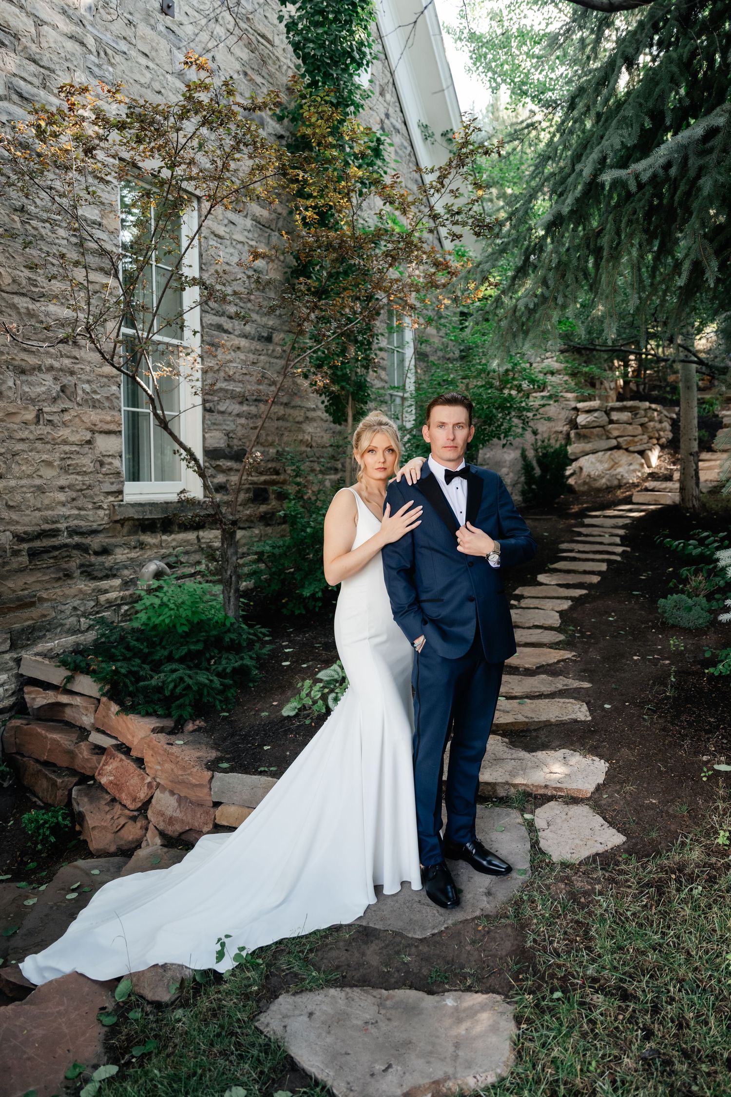 Bride in white mermaid gown embraces groom in navy tuxedo on stone path beside rustic stone of the Washington School House Hotel.