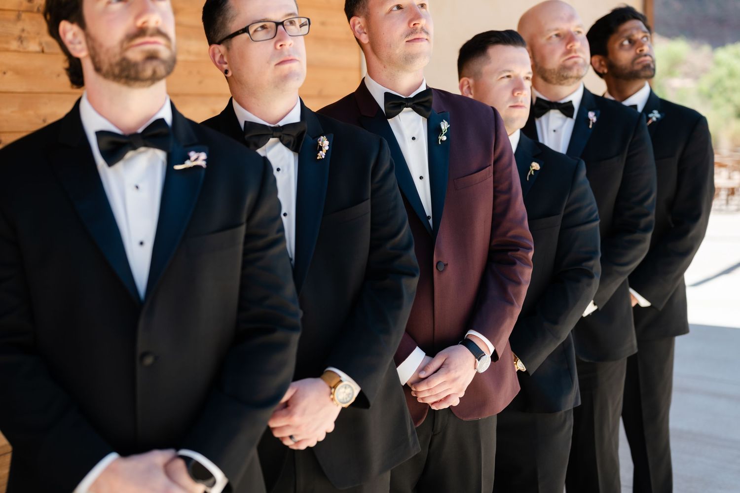 Groom in burgundy tuxedo stands with groomsmen in black tuxedos and bow ties, all wearing boutonnieres.