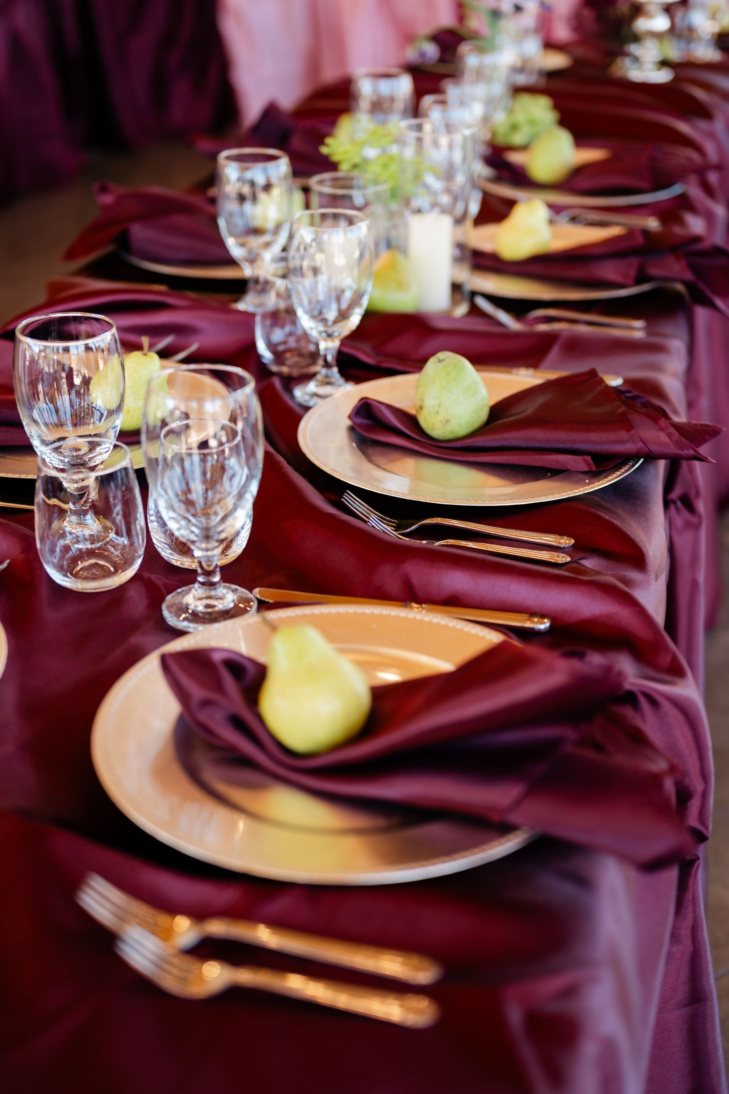 Elegant burgundy table setting with gold plates, crystal glasses, green pears on folded napkins for a formal event.