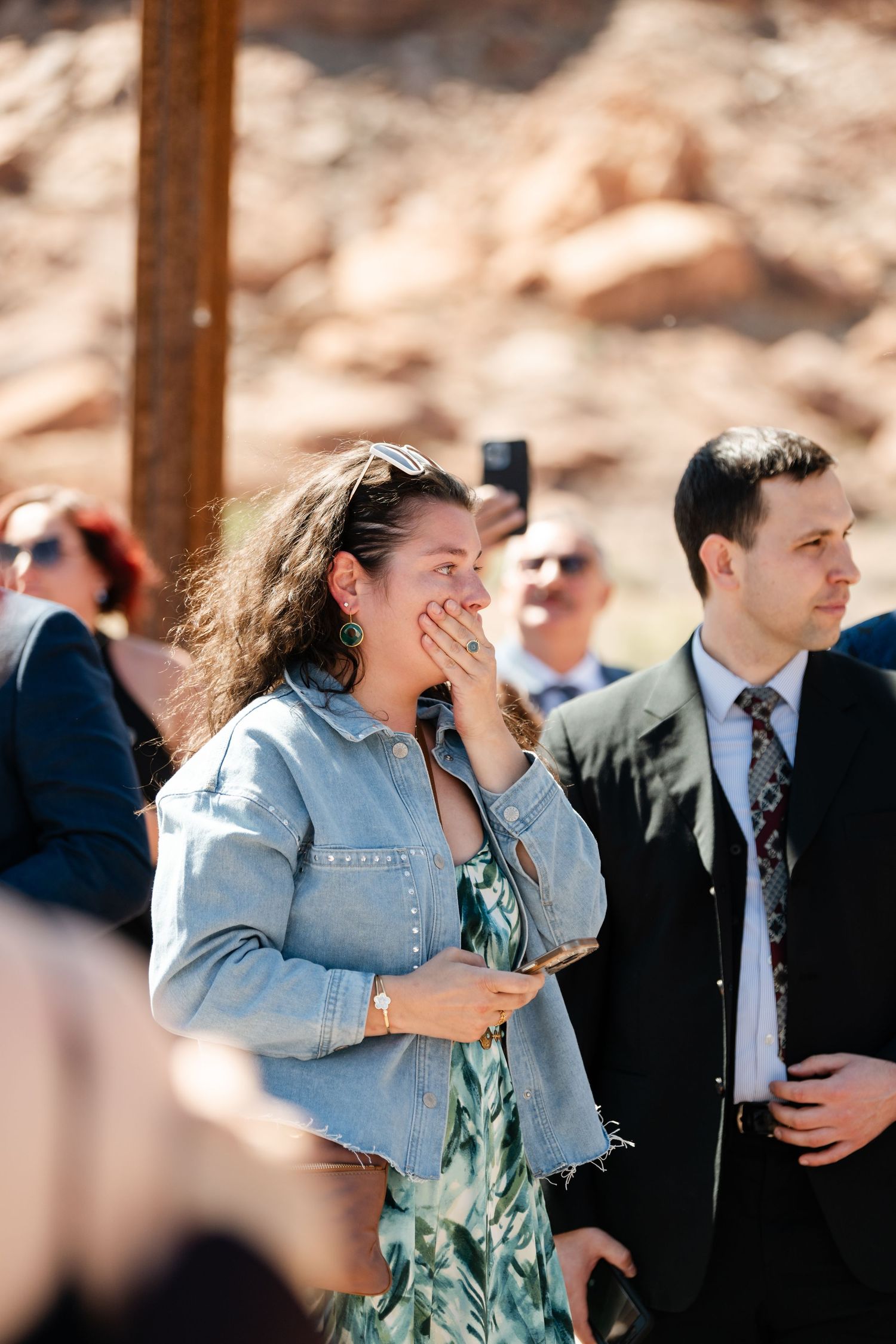 Emotional wedding guest in denim jacket and floral dress covering mouth while watching outdoor ceremony.