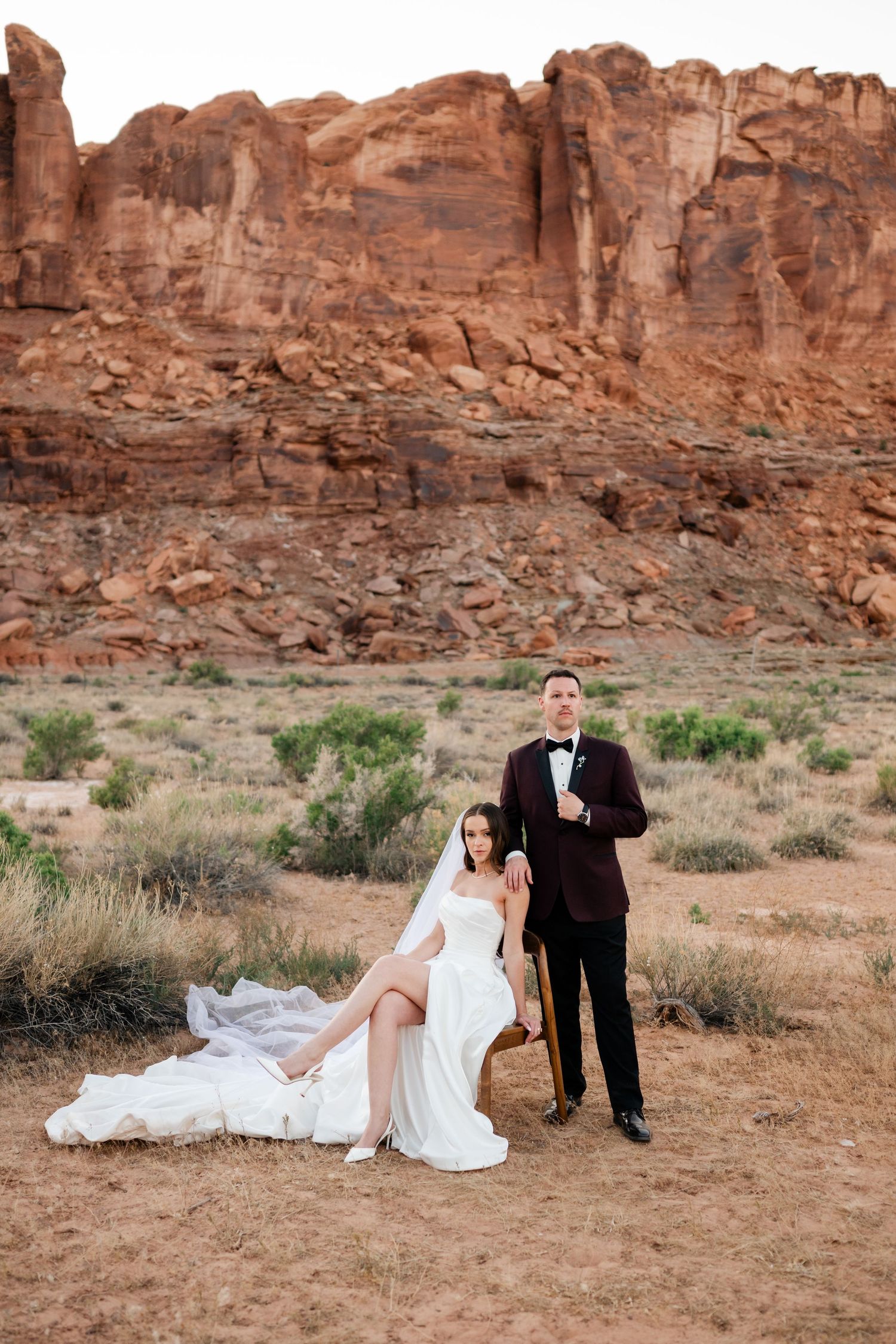 Bride and groom pose before red rock canyon formations in Utah desert at dusk.